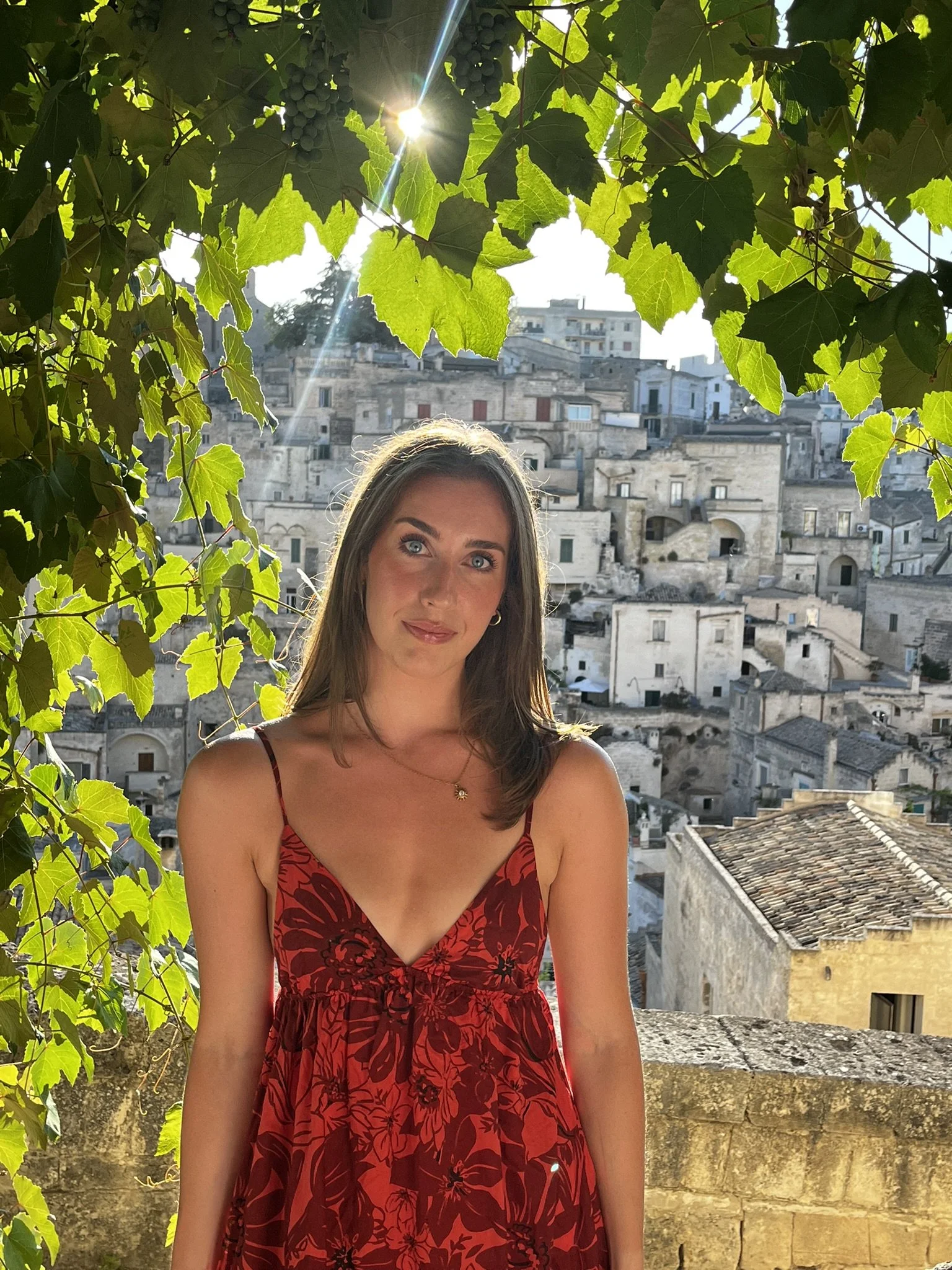 Woman in a red floral dress posing beneath grapevines overlooking Matera's historic stone buildings in Italy. Hand painted surface print designs. Freelance print designer.