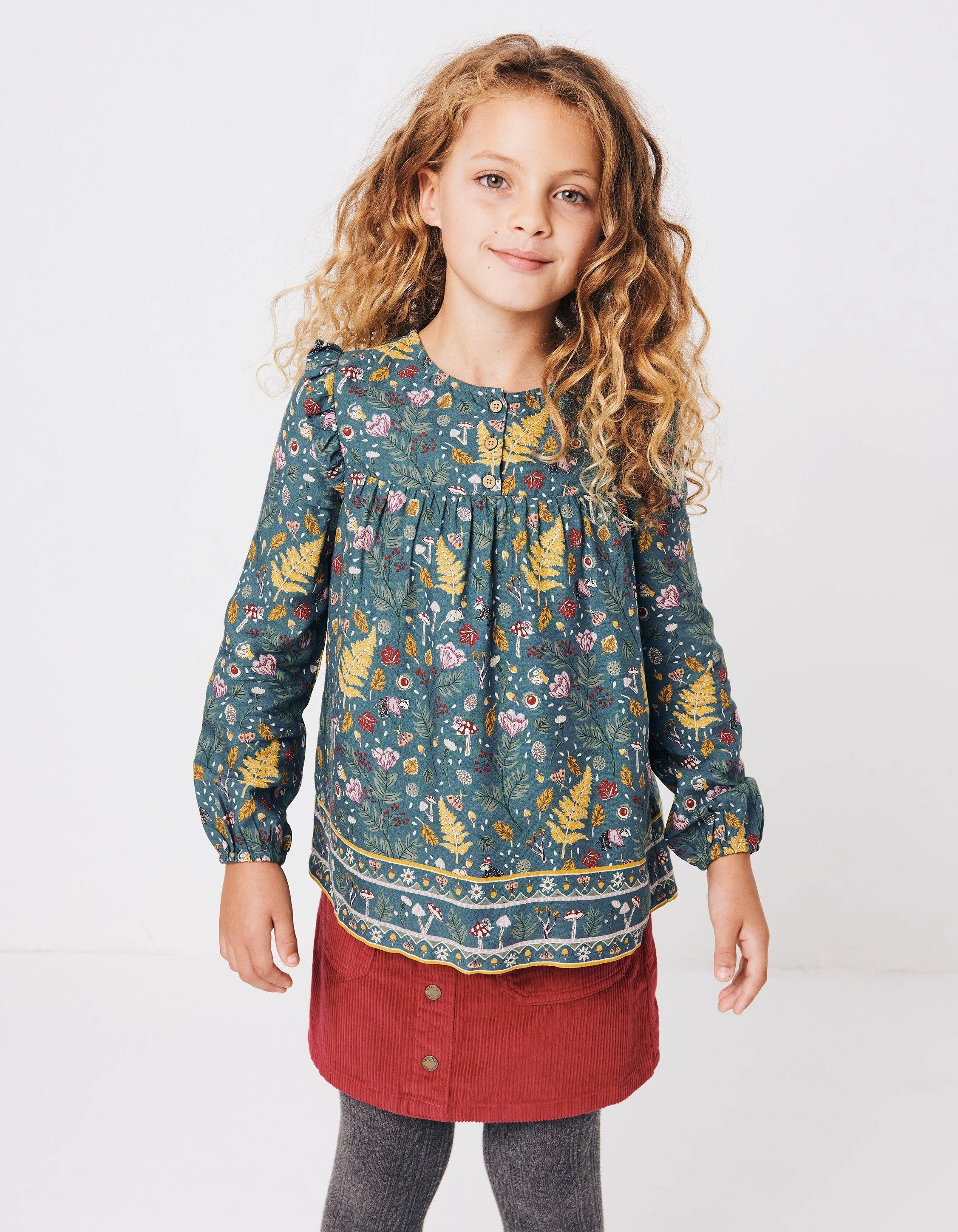 Smiling young girl with curly blonde hair wearing a blue floral-print long-sleeve blouse and red corduroy skirt. Hand painted surface print designs. Freelance print designer.