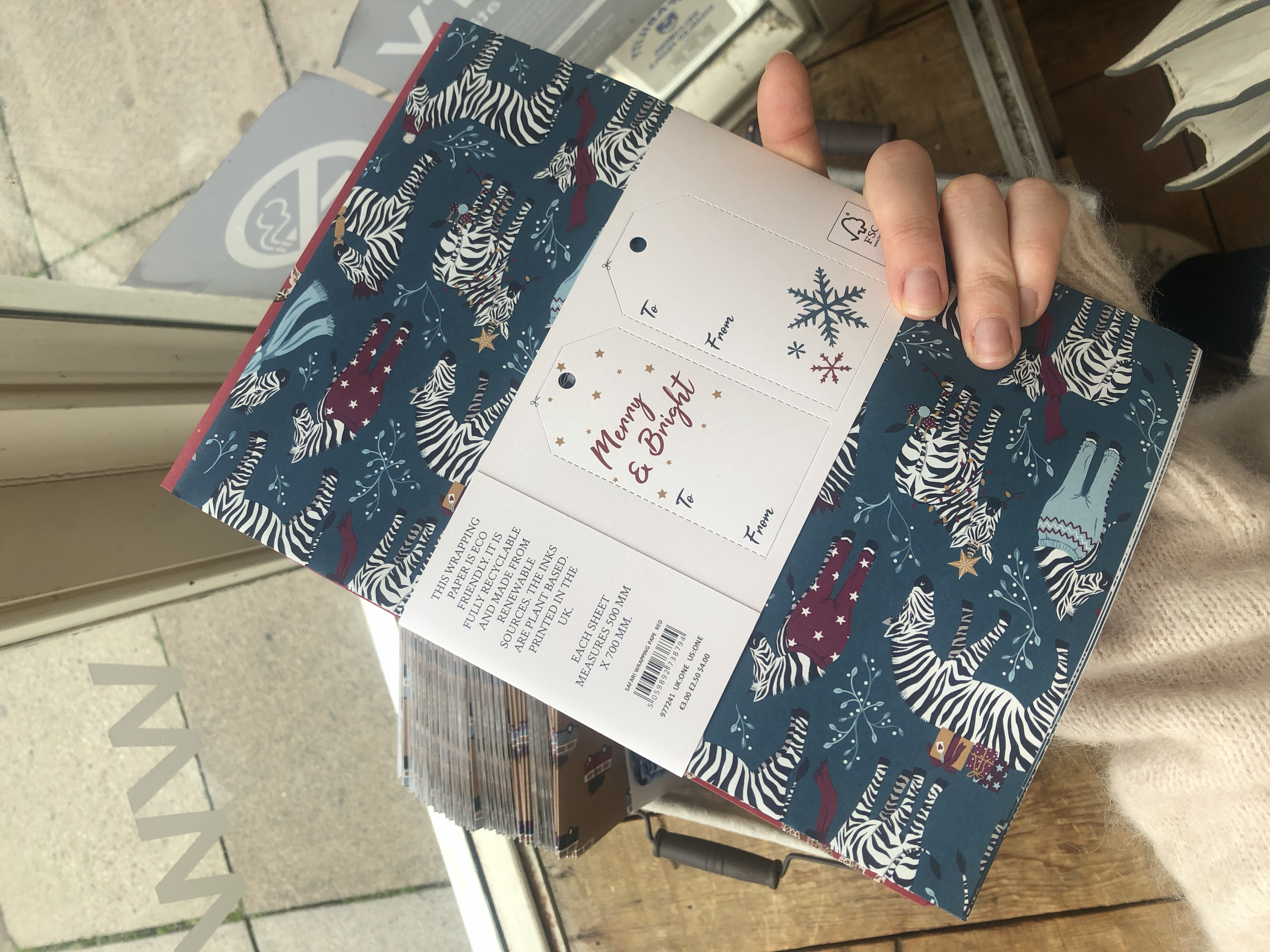 A hand holds eco-friendly Christmas wrapping paper decorated with zebras wearing scarves, hats, and festive clothing, with gift tags and snowflakes. Hand painted surface print designs. Freelance print designer.