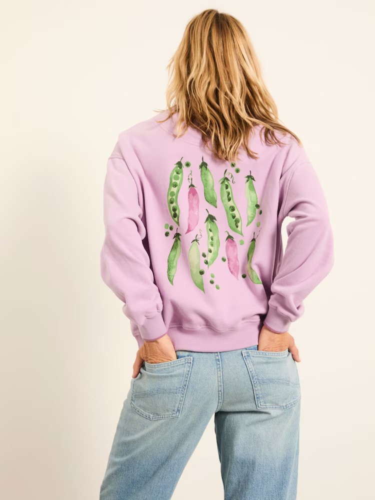 Back view of woman wearing a pink sweatshirt with colorful pea pod artwork and light blue jeans. Hand painted surface print designs. Freelance print designer.