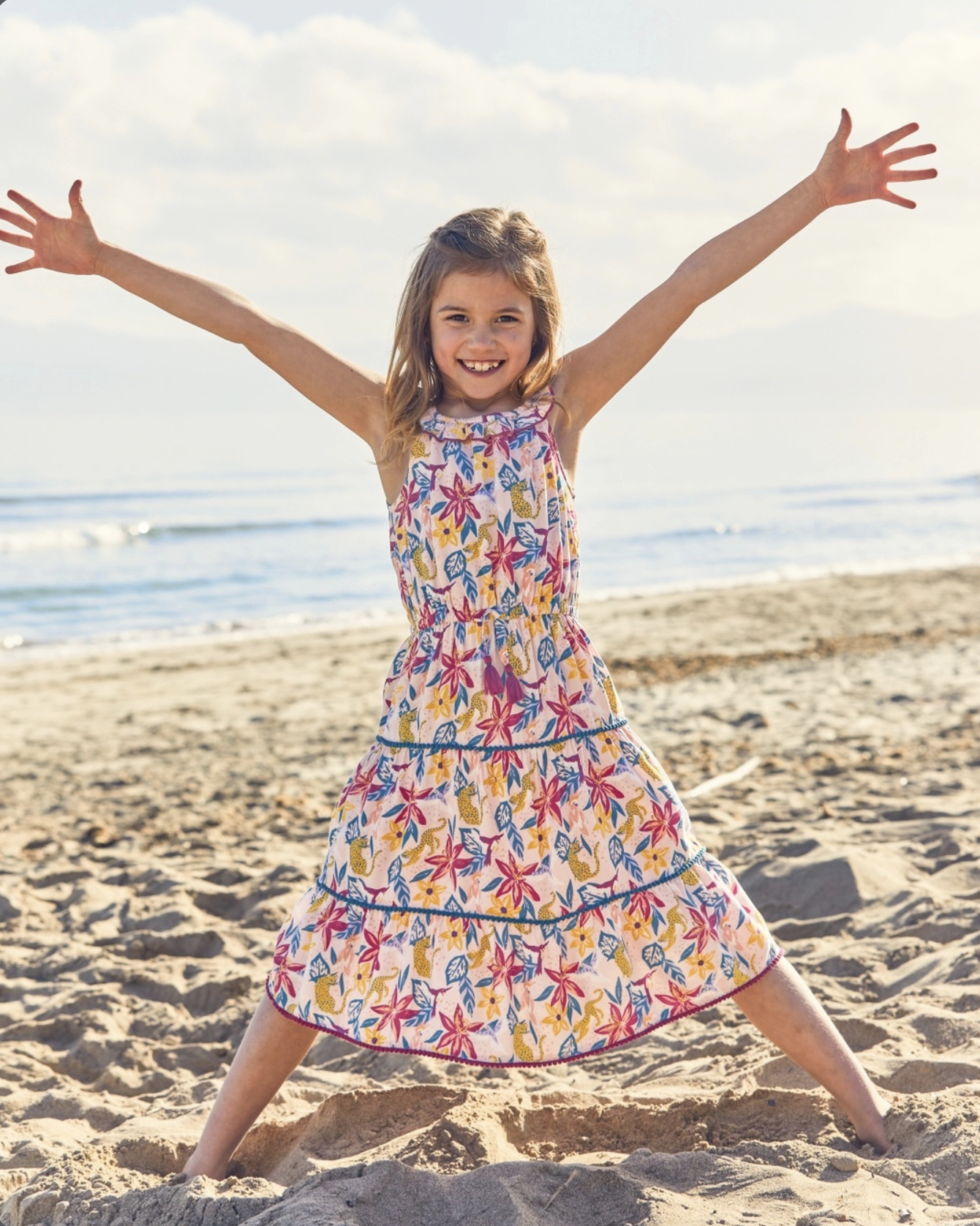 Smiling young girl in a colorful floral dress standing with arms outstretched on a sunny beach. Hand painted surface print designs. Freelance print designer.