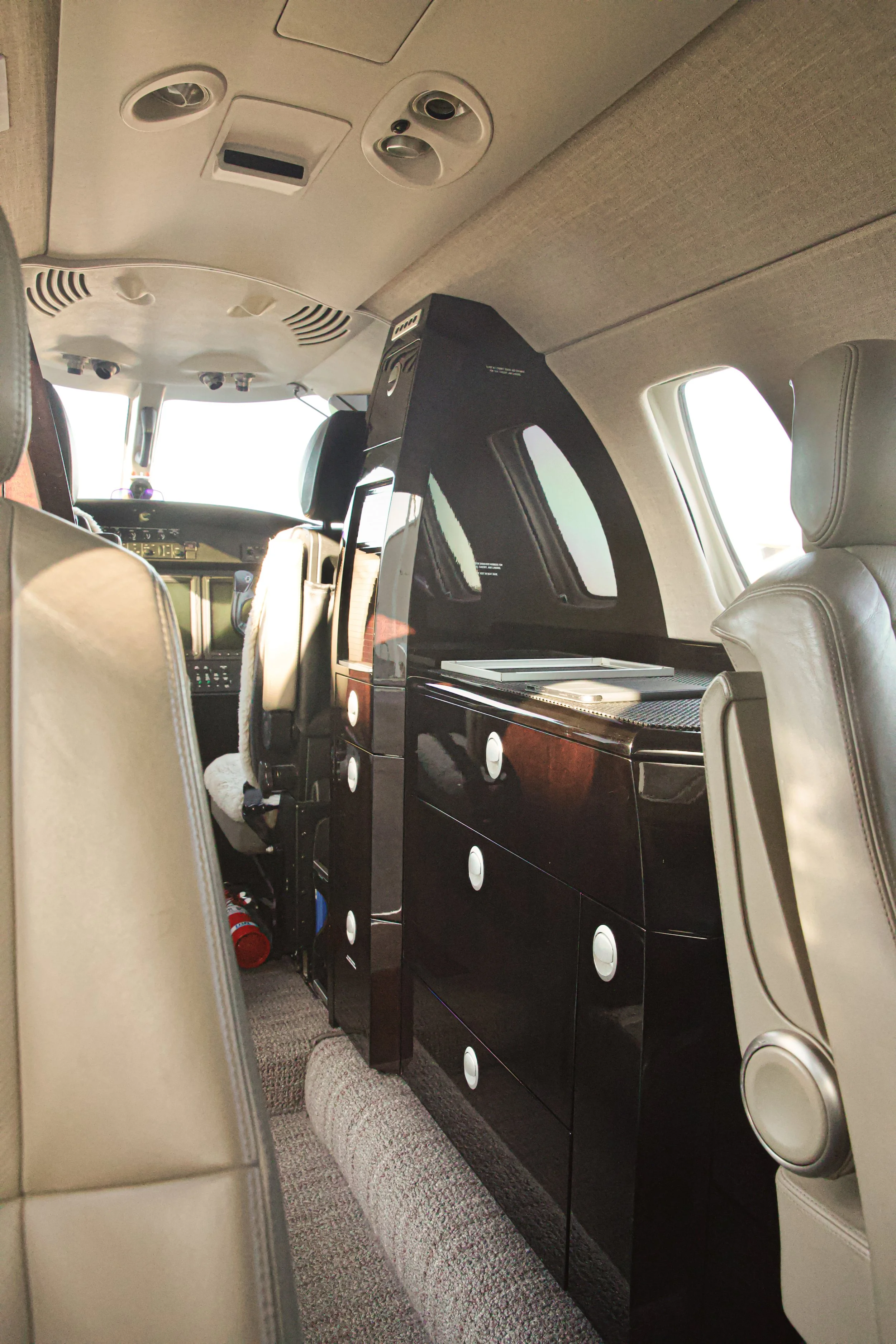 Interior of a small aircraft showing overhead controls, cockpit, passenger seats, and a black equipment cabinet.