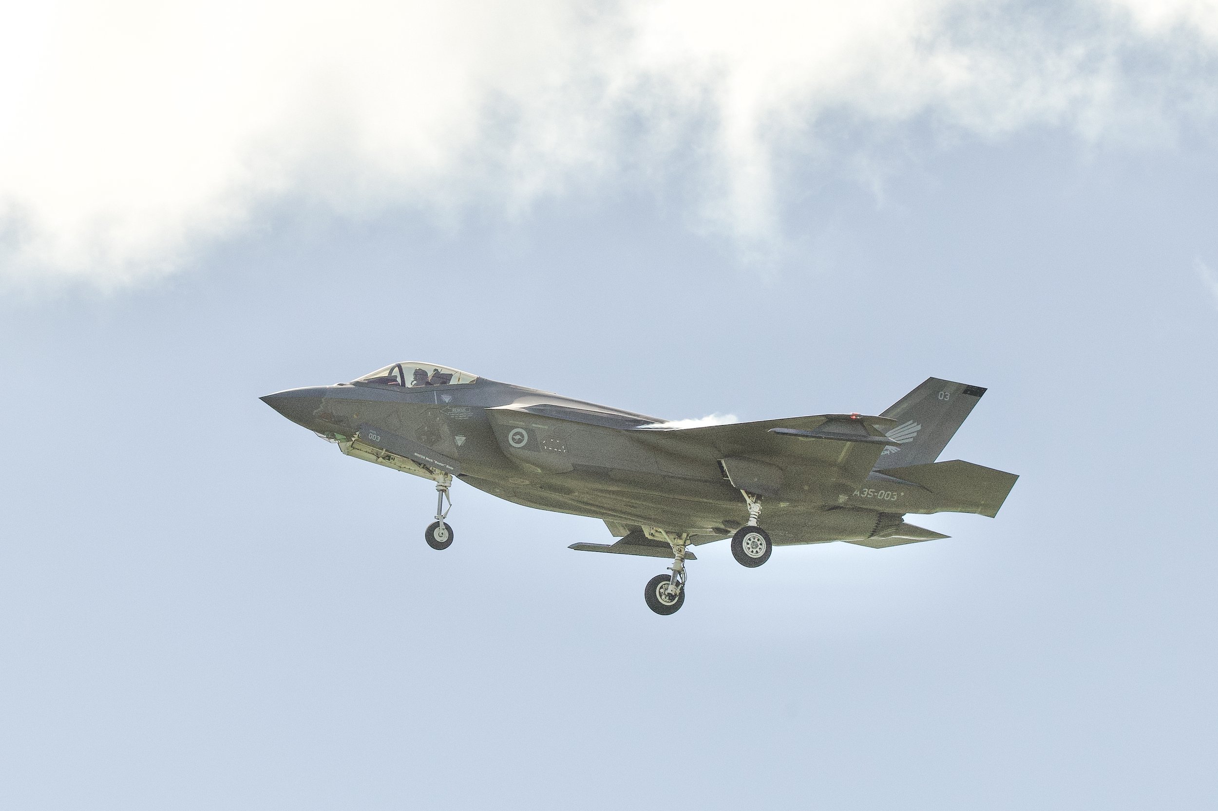 F-35 Lightning II fighter jet flying with landing gear extended against a cloudy sky
