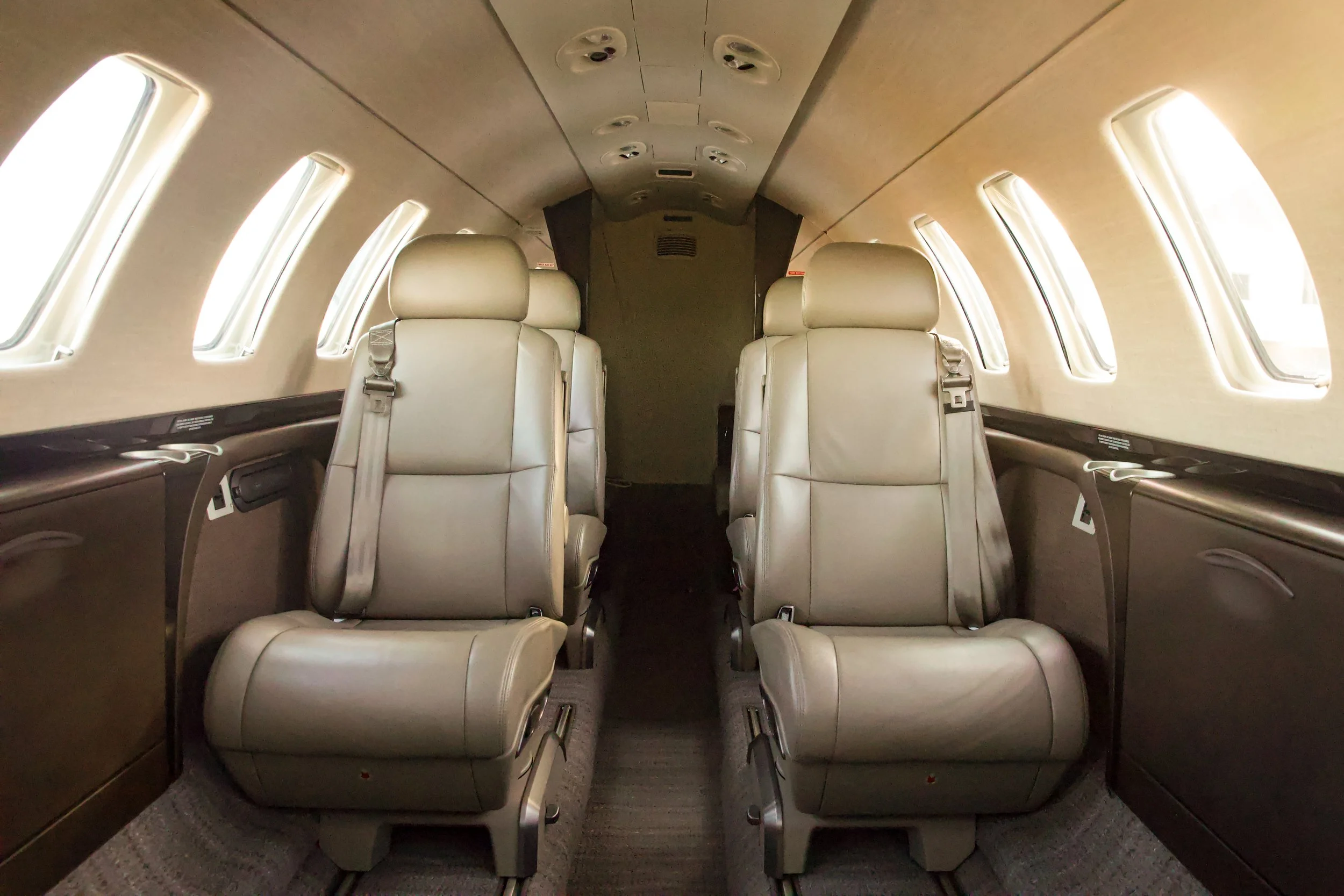 Interior of a small private jet with four cream leather seats, seat belts, oval windows, and overhead air vents.