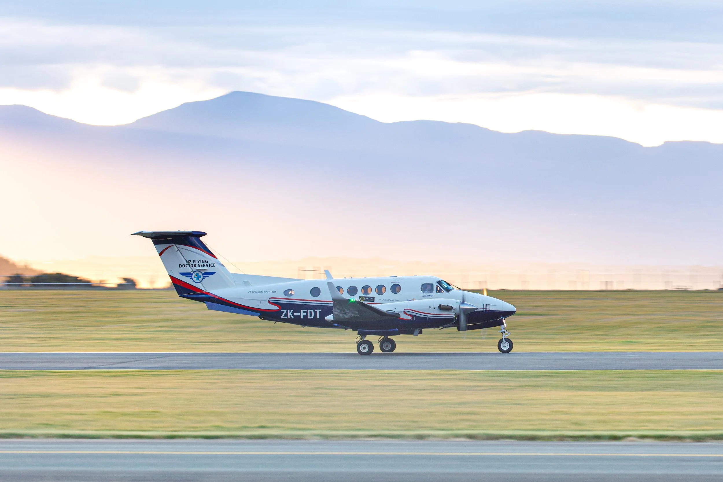 NZ Flying Doctor Service twin-engine aircraft taxiing on a runway at sunset with mountains in the background