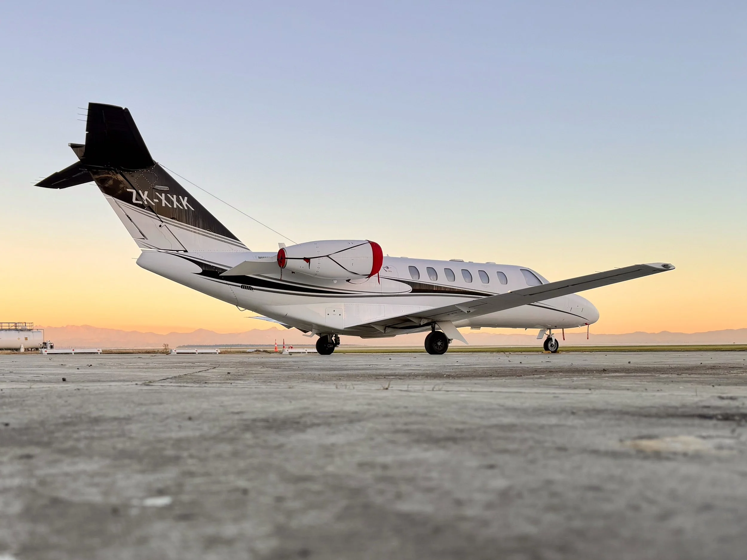 White private jet parked on a runway at sunset, viewed from the rear side with mountains in the background.