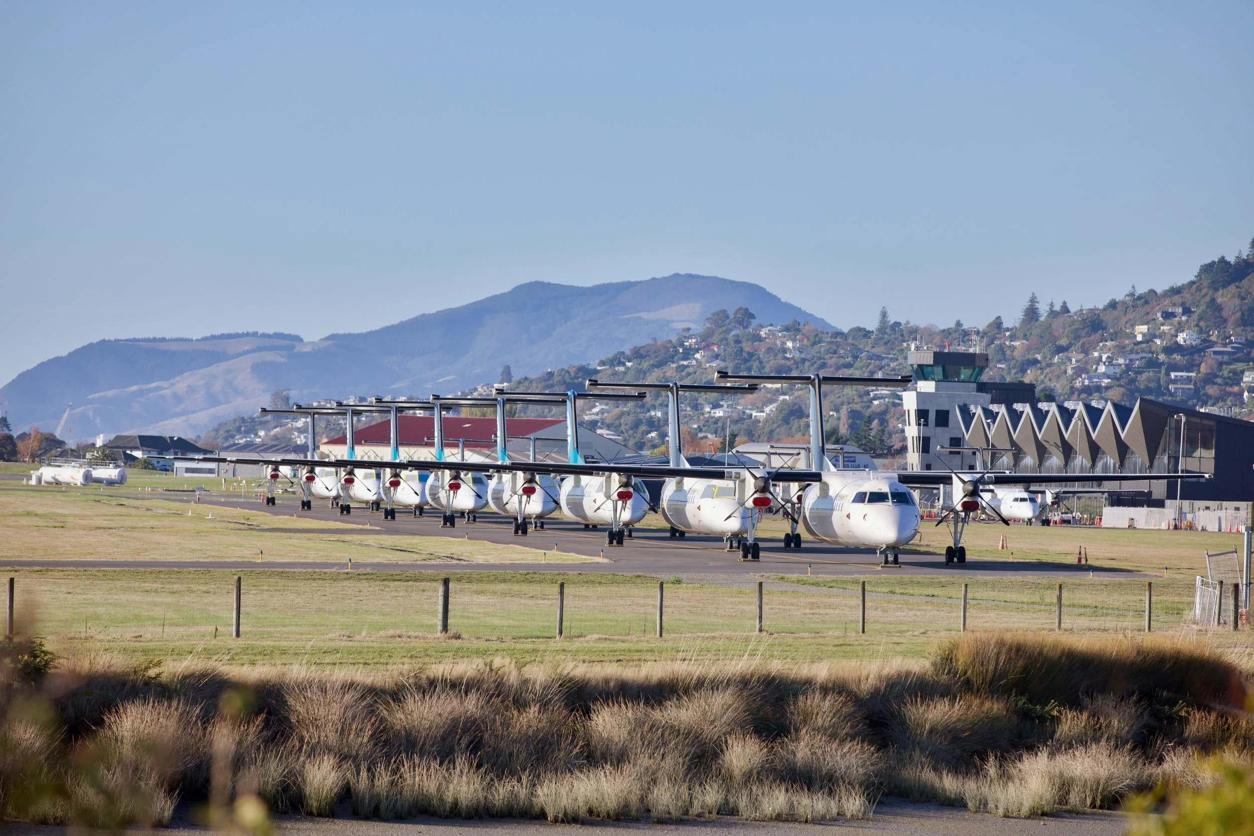 Several turboprop passenger planes parked at an airport with a control tower, hillside homes, and mountains in the background