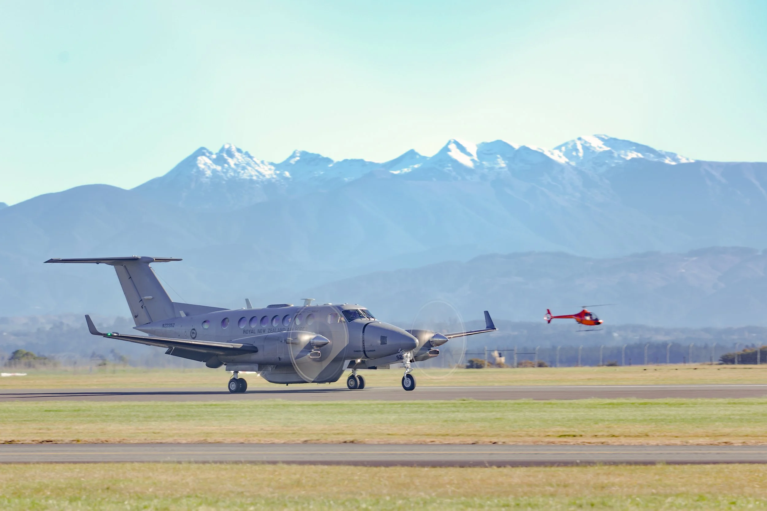 Royal New Zealand Air Force twin-engine aircraft taxiing on a runway with a red helicopter and snow-capped mountains in the background