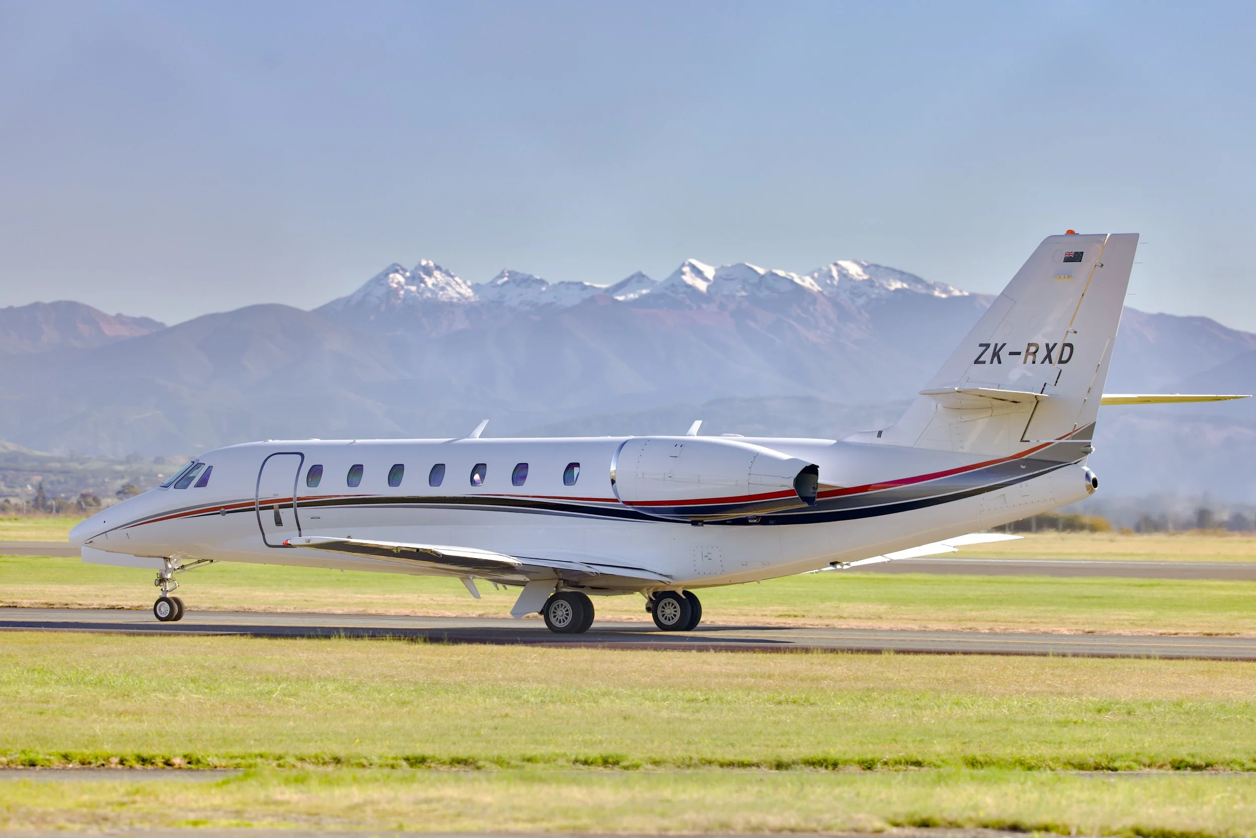 White private jet with red and black stripes on a runway, with snow-capped mountains in the background