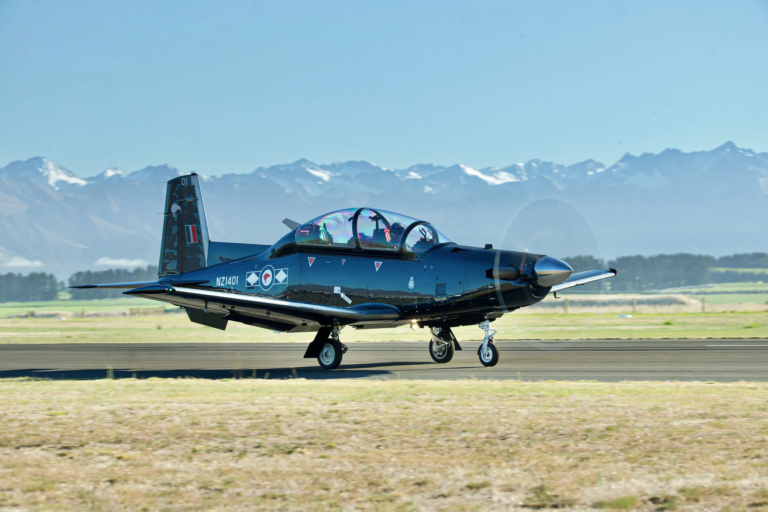 Dark blue single-engine military trainer aircraft taxiing on a runway with snow-capped mountains in the background