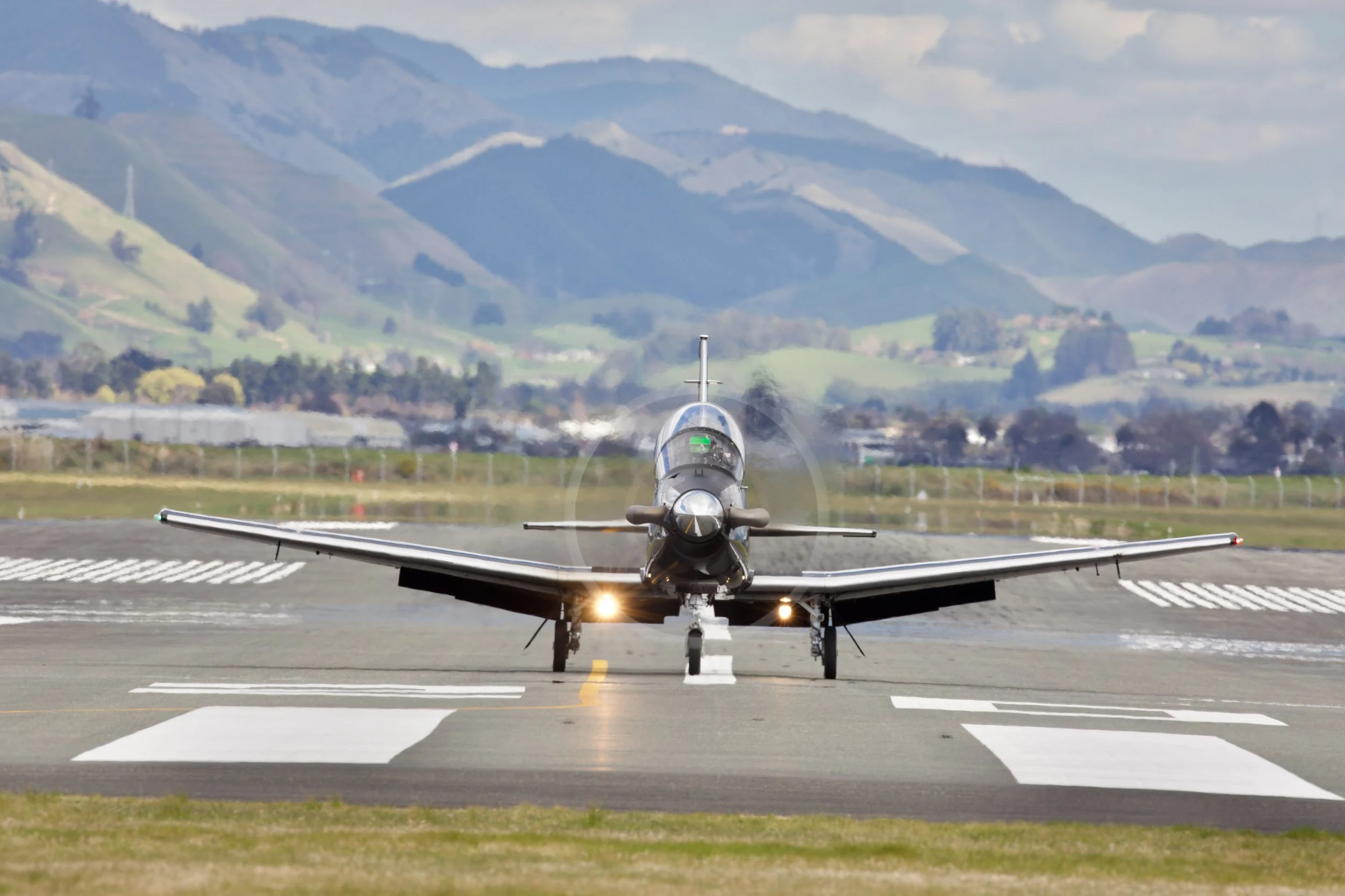 Front view of a small propeller aircraft taxiing on a runway with mountains in the background