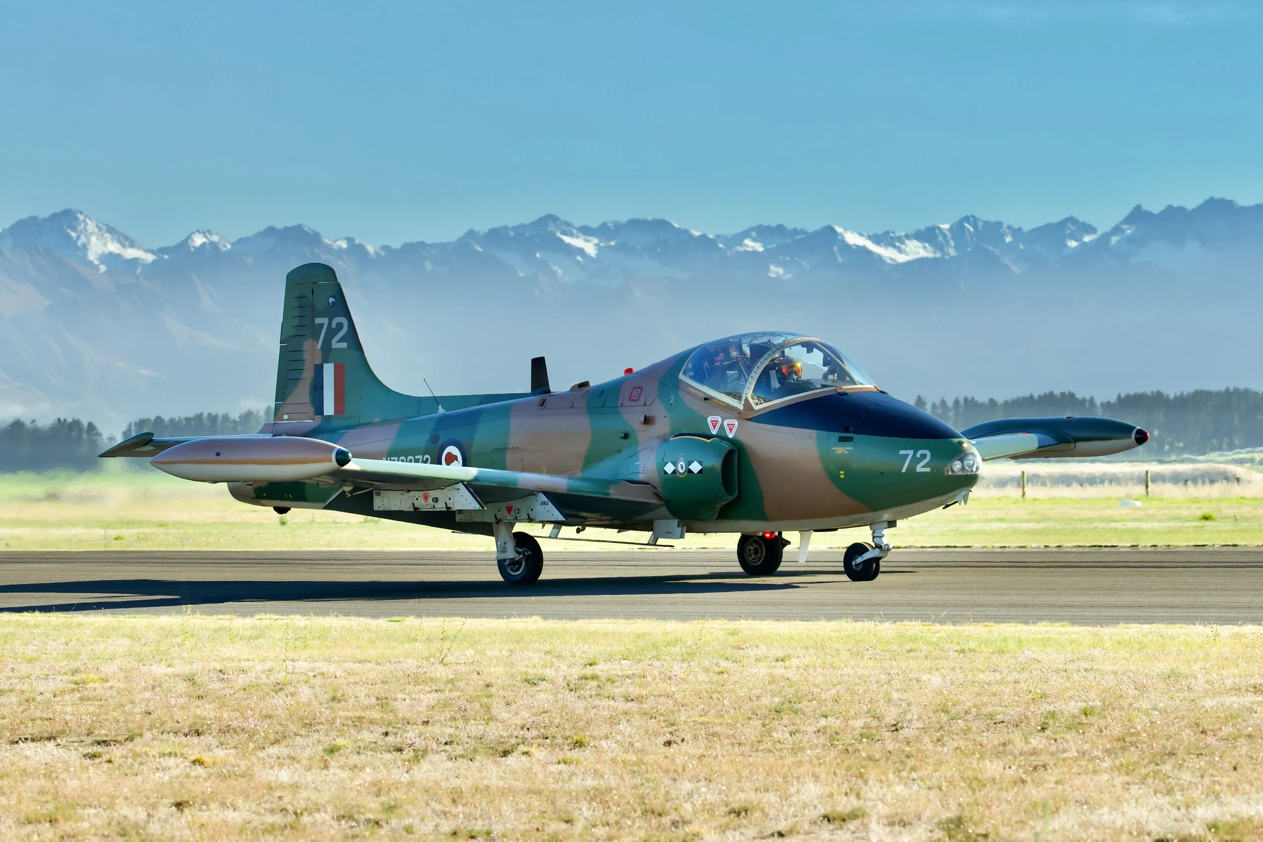 Camouflaged military jet marked 72 on a runway with snow-capped mountains in the background