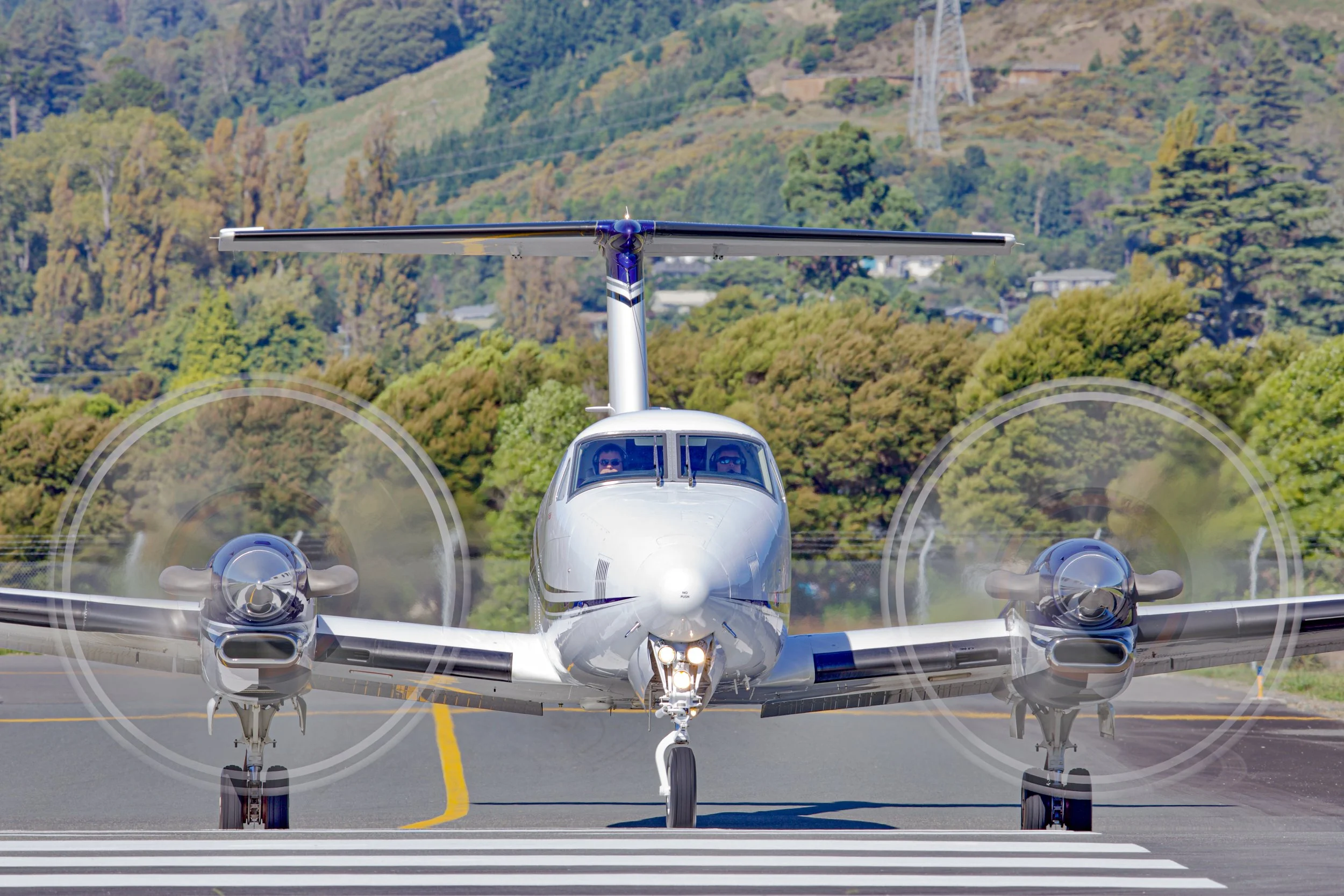 Front view of a twin-engine propeller airplane on a runway with two pilots visible in the cockpit