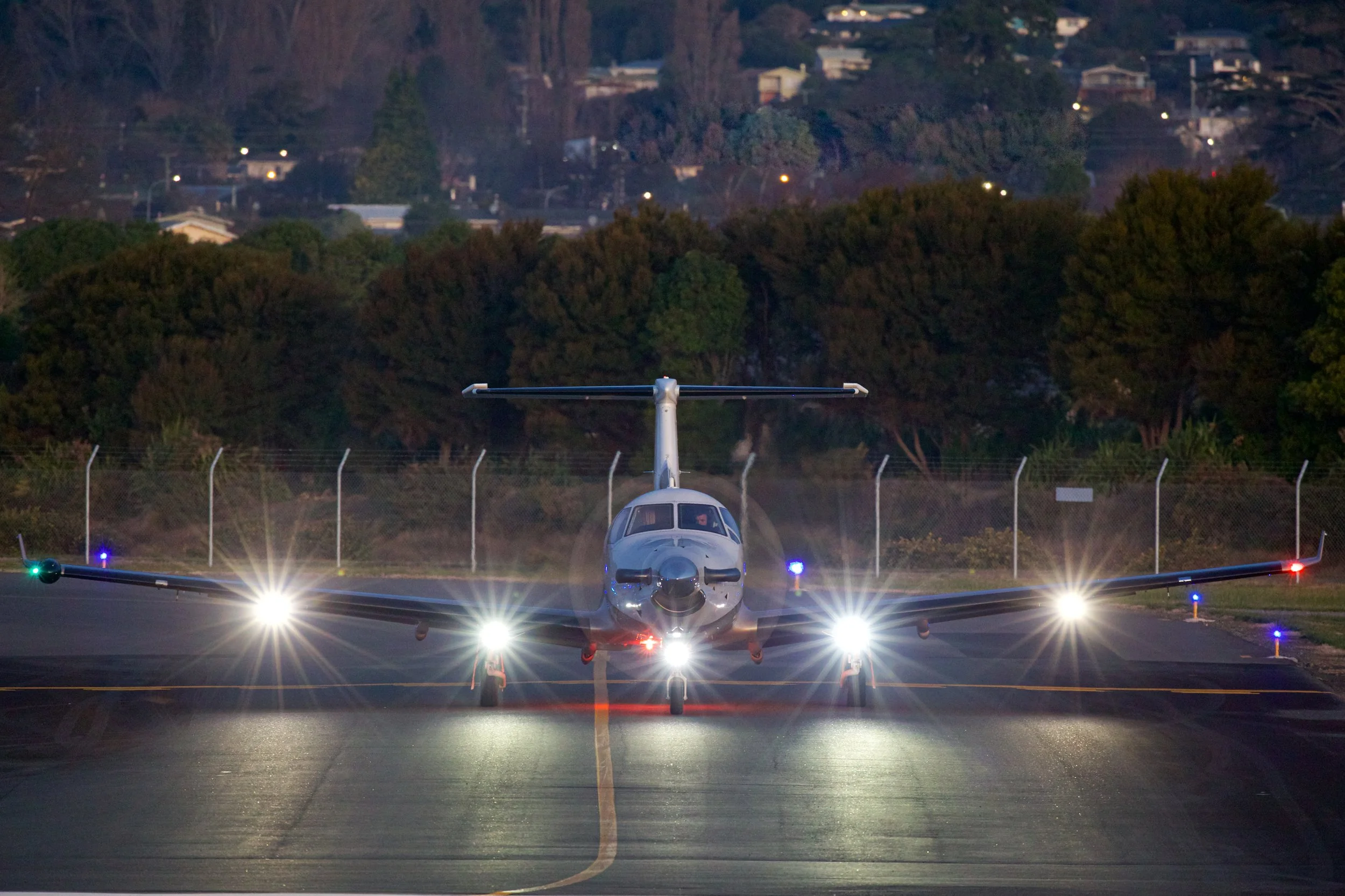Twin-engine propeller airplane taxiing on a runway at dusk with landing lights illuminated
