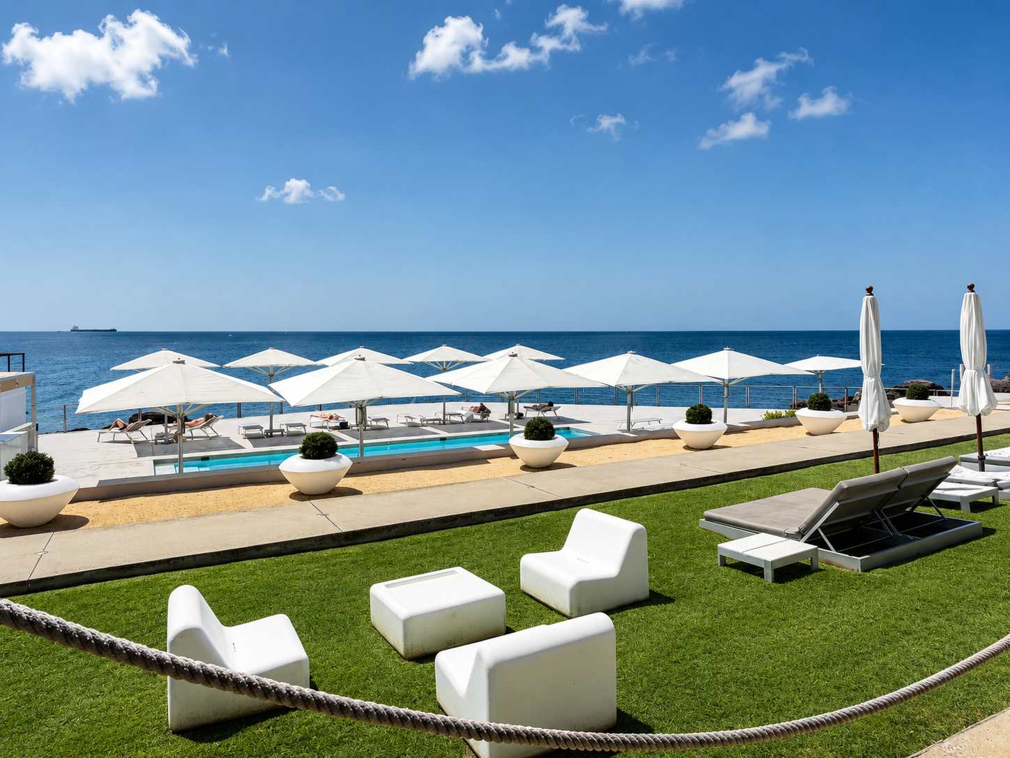 Oceanfront resort pool deck with white umbrellas, lounge chairs, manicured lawn, and blue sea under a sunny sky