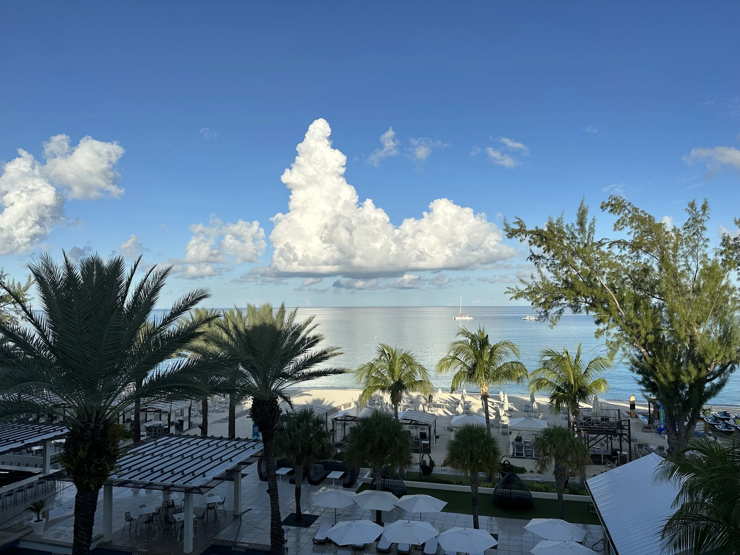 Tropical beach resort overlooking calm turquoise water with palm trees, lounge umbrellas, sailboats, and a bright blue sky