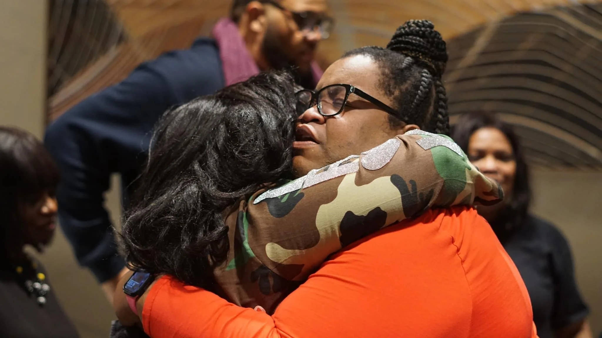 Two women embracing in a crowded indoor gathering, with onlookers in the background.