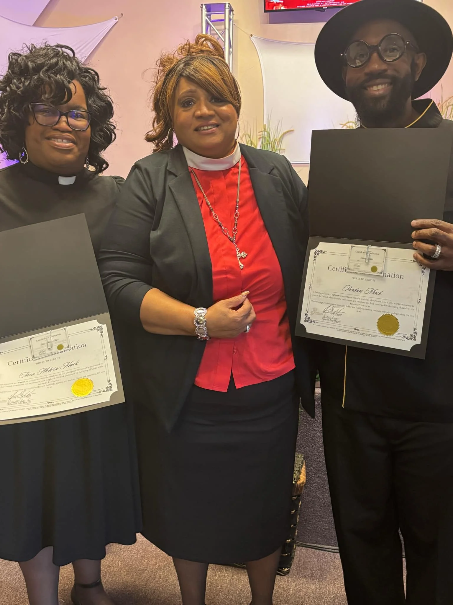 Three people pose at a church ceremony holding framed ordination certificates, including two clergy members and a man in a black hat.