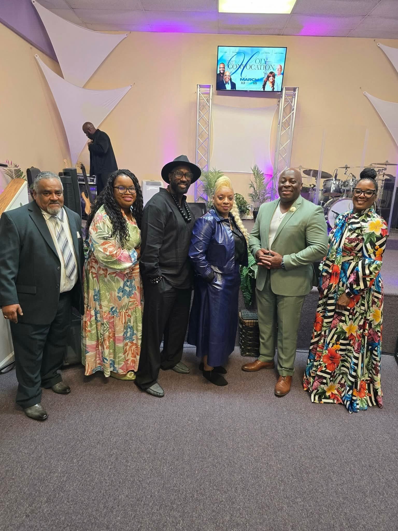 Six adults pose for a group photo on a church stage with musical instruments and a concert announcement screen behind them.