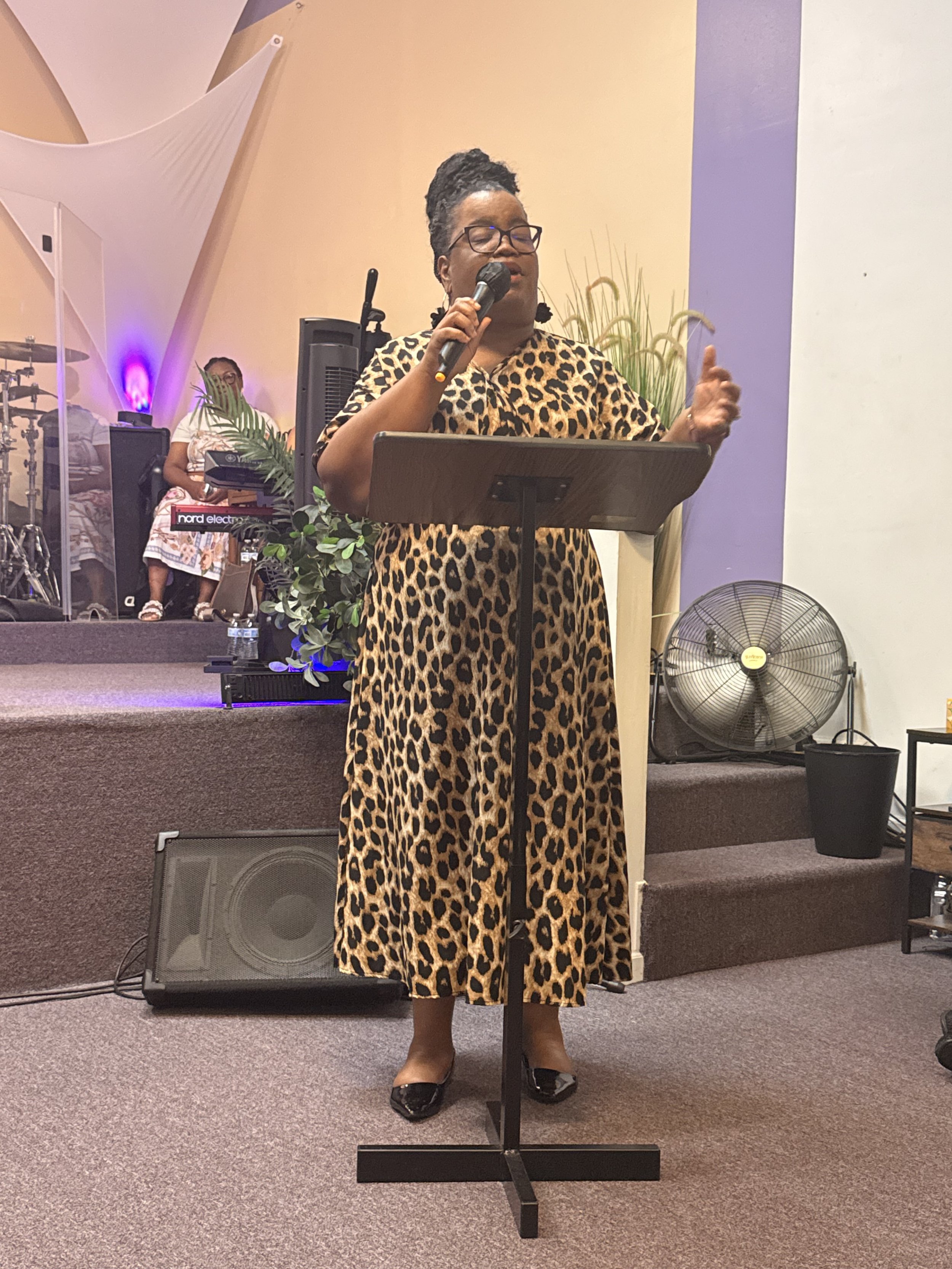 Woman in a leopard-print dress singing into a microphone behind a music stand on a church stage.