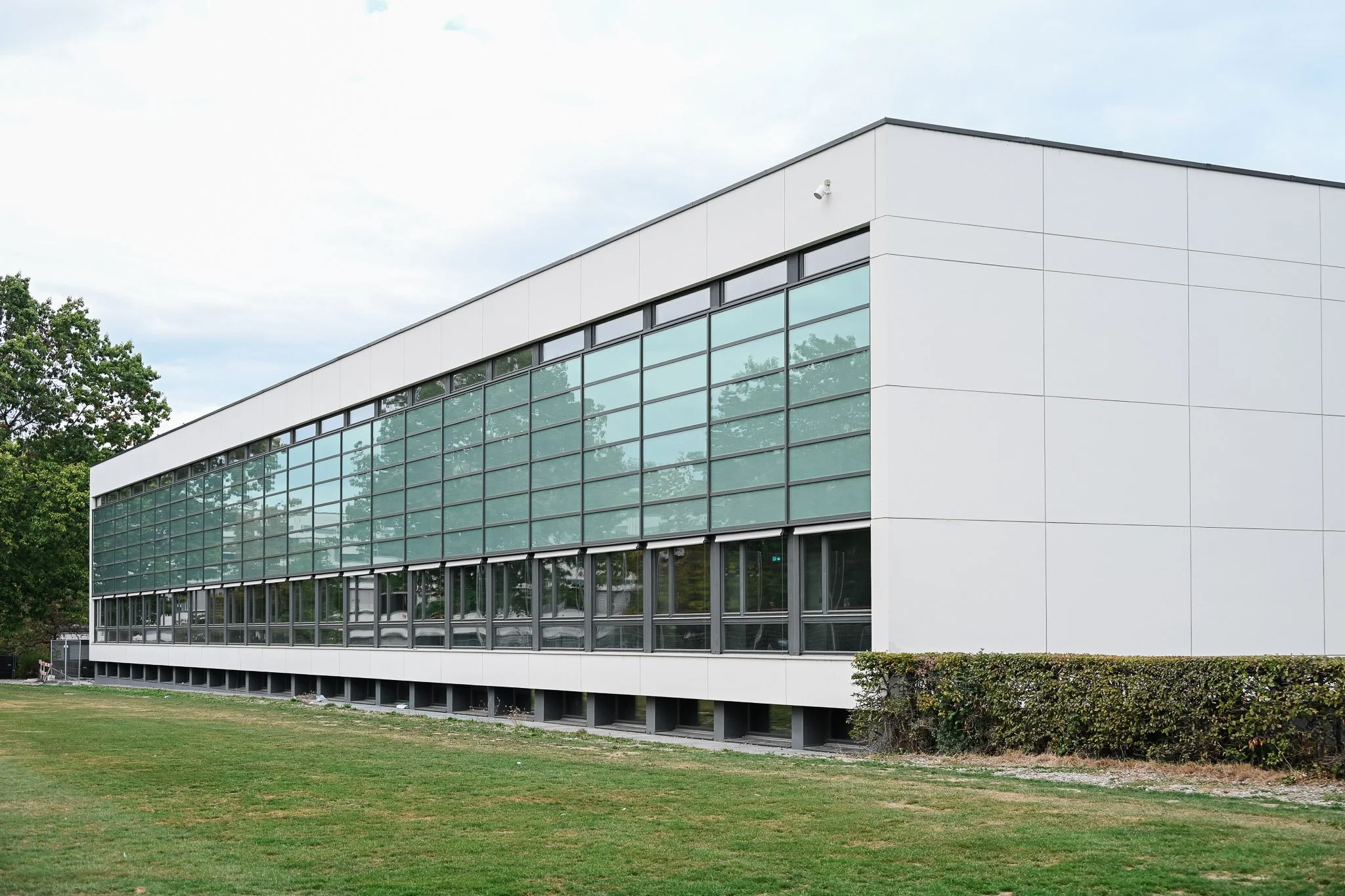 Modernes, weiß verkleidetes Gebäude mit großer Glasfassade, Rasenfläche und Hecke im Vordergrund