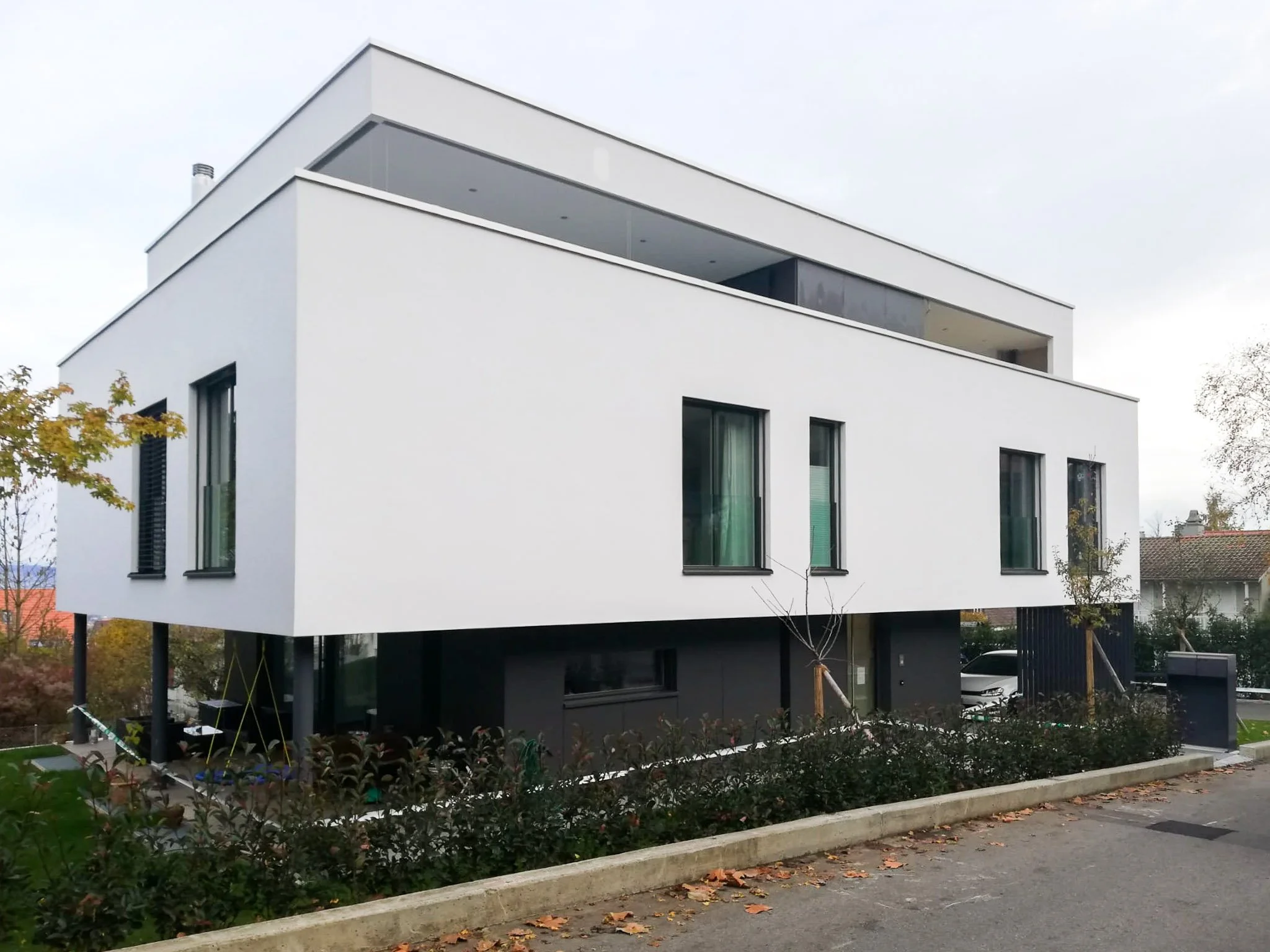 Modernes weißes Einfamilienhaus mit Flachdach, dunklen Fenstern, überdachter Terrasse und gepflegtem Vorgarten