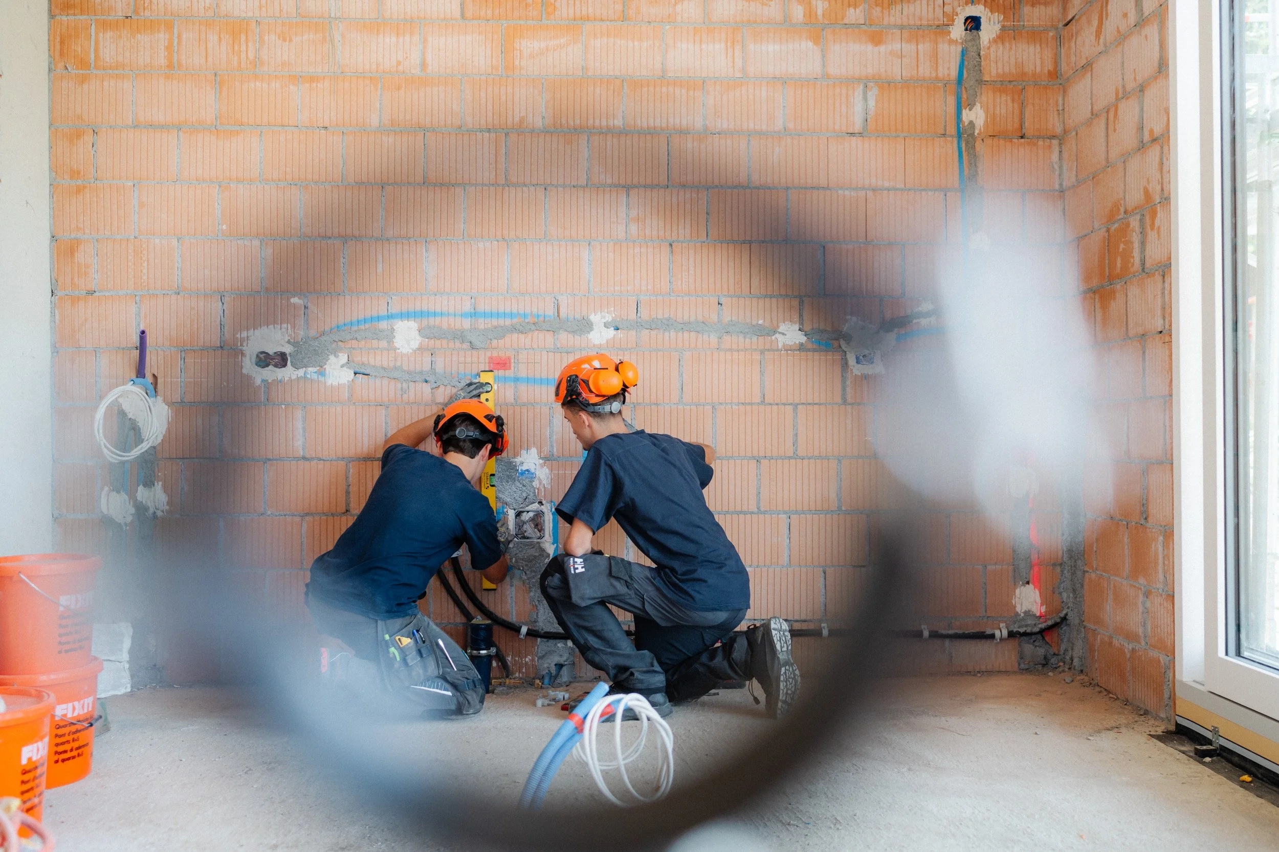 Zwei Handwerker mit Schutzhelmen installieren Leitungen in einem Rohbau mit unverputzten Ziegelwänden.