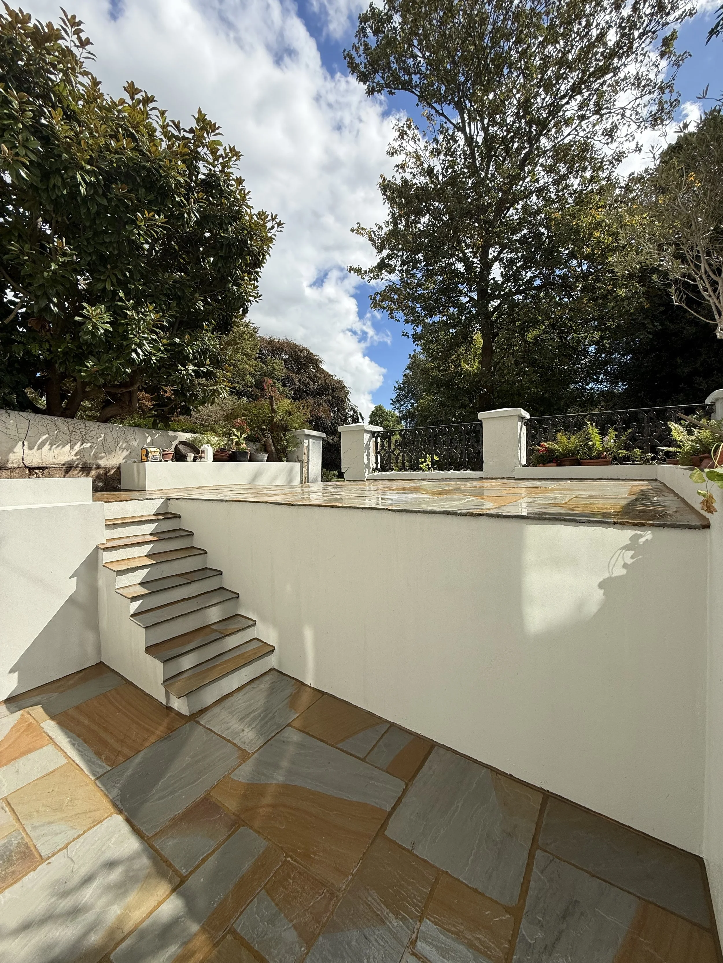 Sunlit garden patio with slate paving, white walls and steps, black railing, potted plants, and leafy trees beneath a partly cloudy blue sky