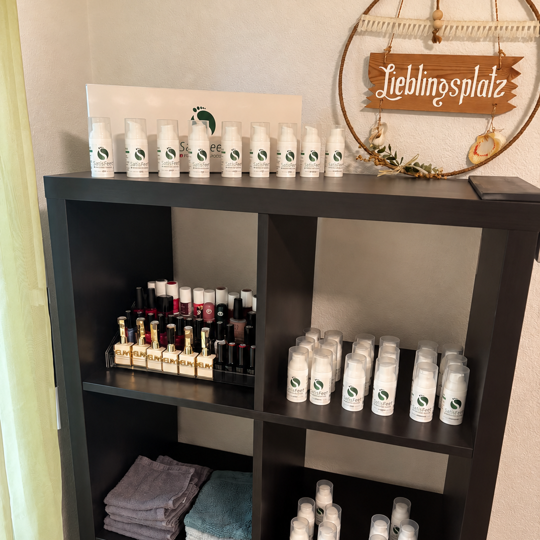 Schwarzes Regal mit SatisFeet-Produkten, Nagellacken, Handtüchern und dekorativem Holzschild „Lieblingsplatz“