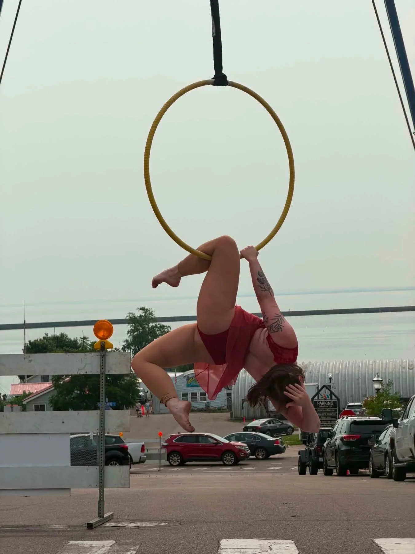 Aerial performer in a red costume hangs upside down from a suspended hoop above a waterfront street.