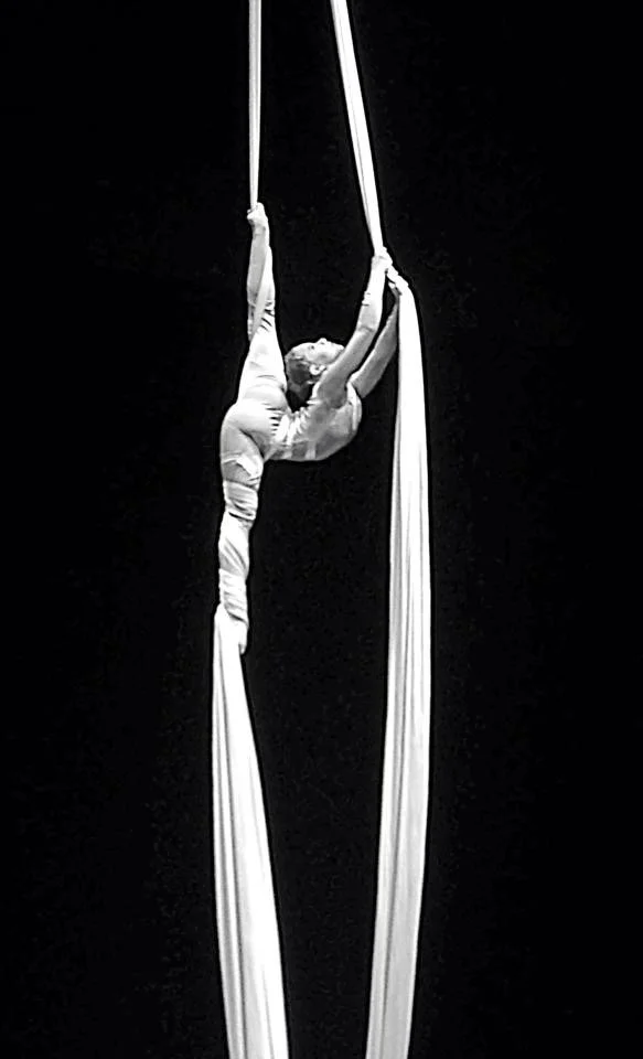 Aerial performer suspended in white aerial silks against a black background.