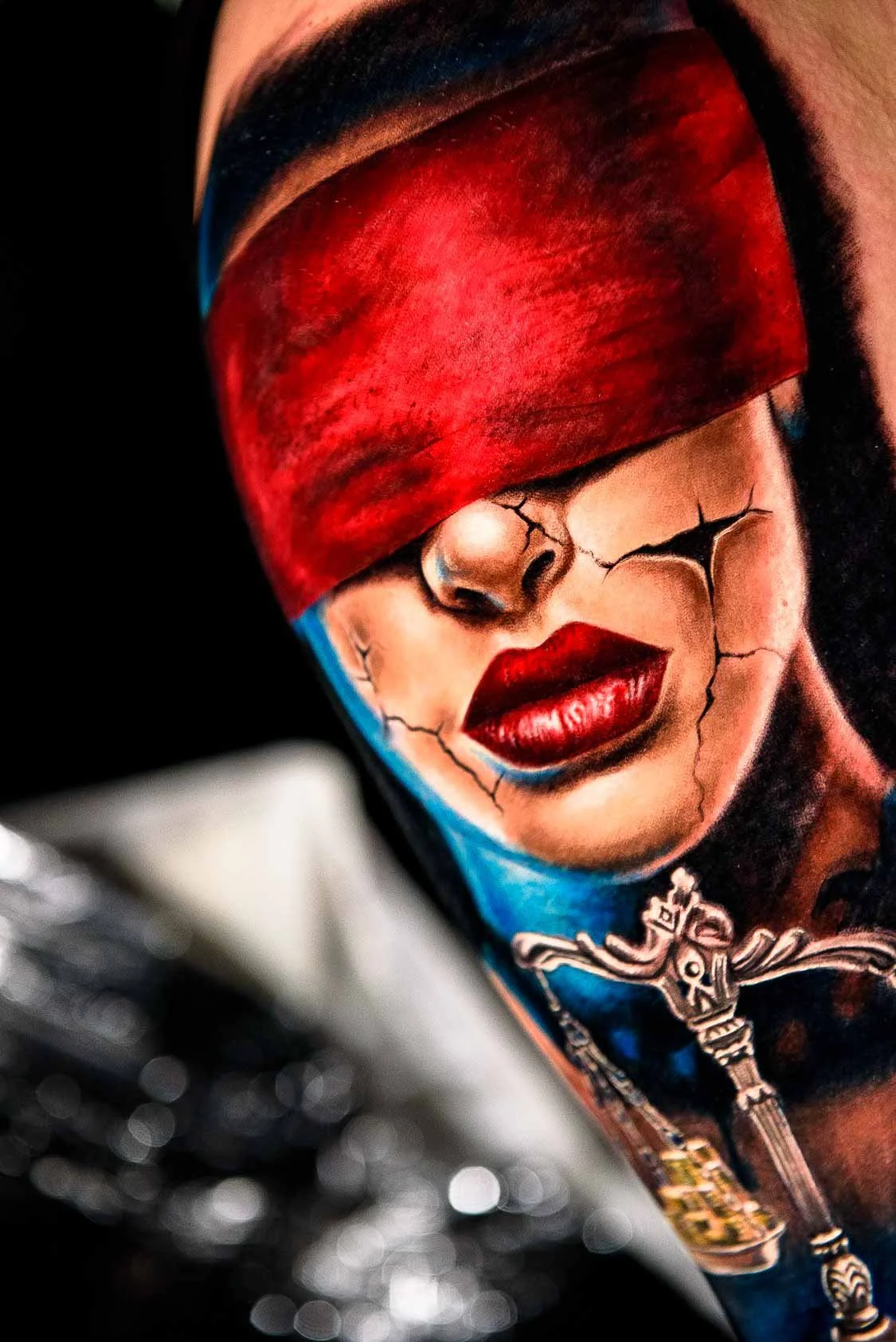Lady Justice tattoo with a red blindfold, red lips, a cracked face and scales against blue tones.