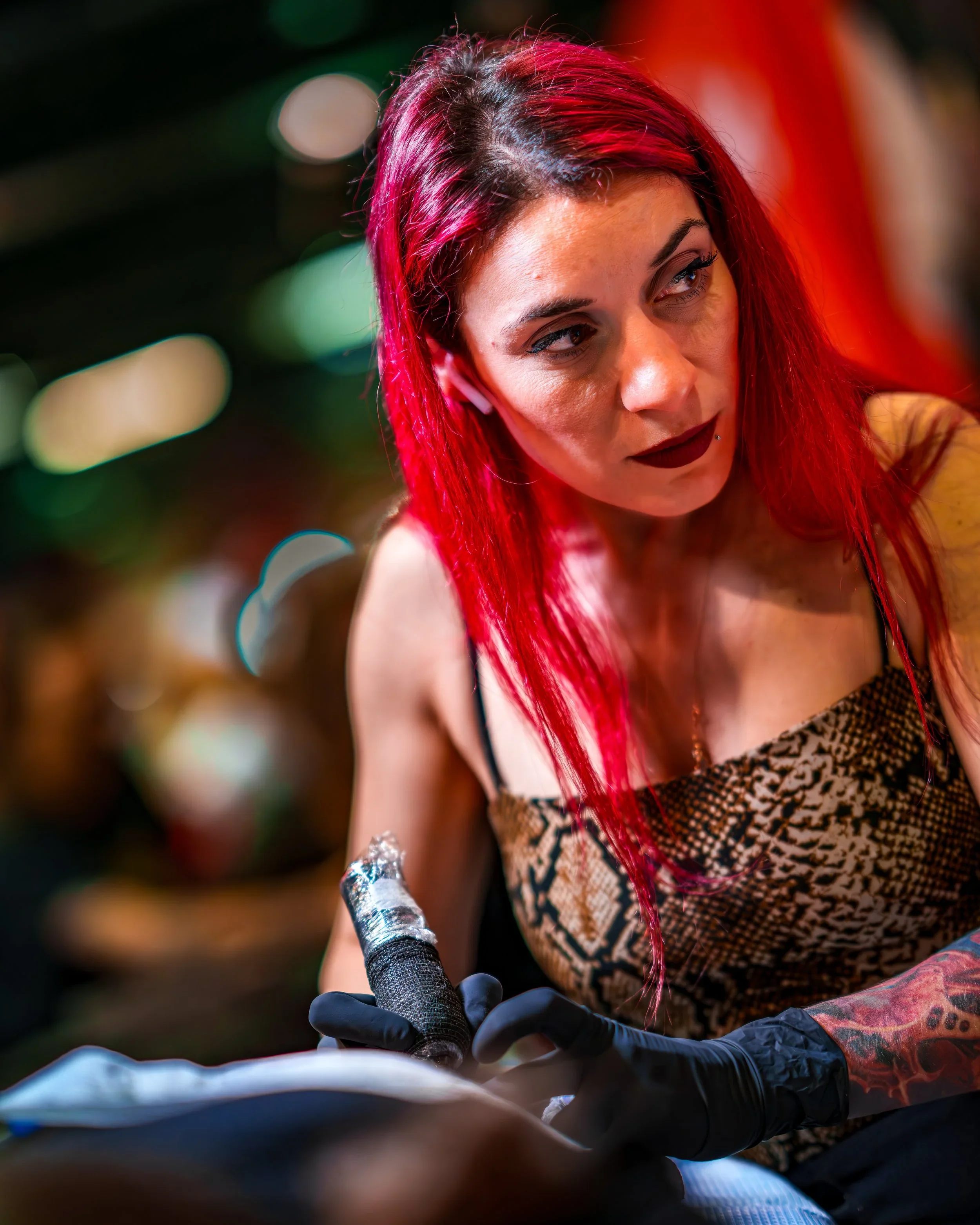 Lena Diamanti with red hair, holding a tattoo machine and wearing black gloves.