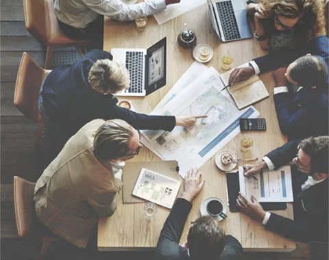 Overhead view of a diverse business team collaborating around a conference table with laptops, blueprints, documents, and coffee.