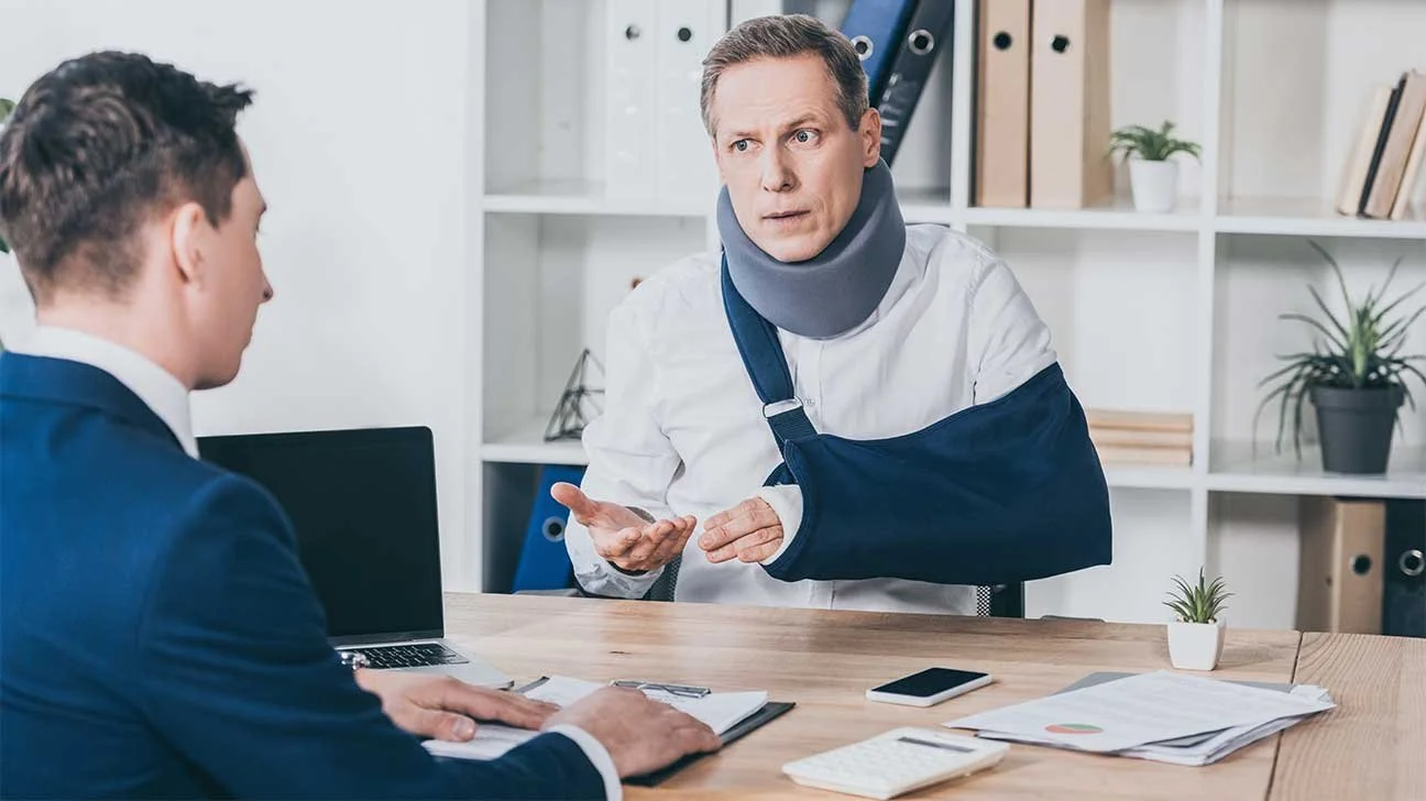Injured businessman wearing a neck brace and arm sling talks with an advisor across a desk