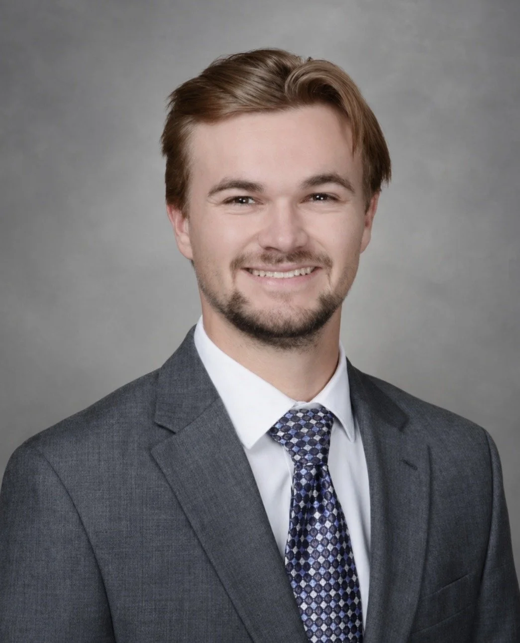 Professional headshot of a smiling man in a gray suit, white shirt, and patterned tie against a gray background