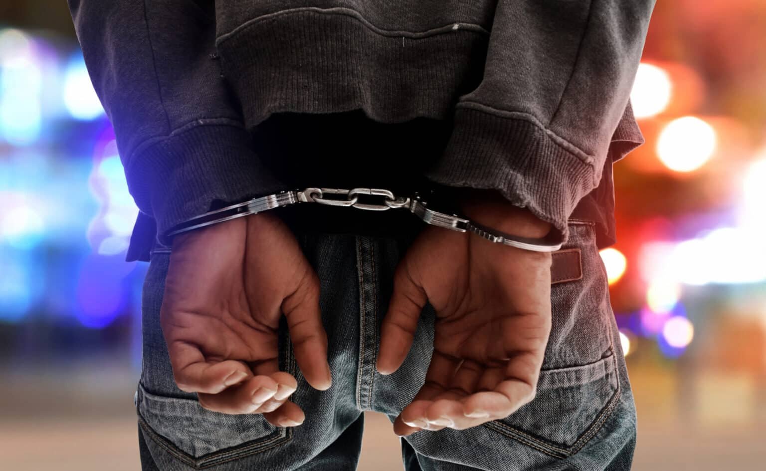Person in handcuffs with hands behind their back, with blurred police lights in the background