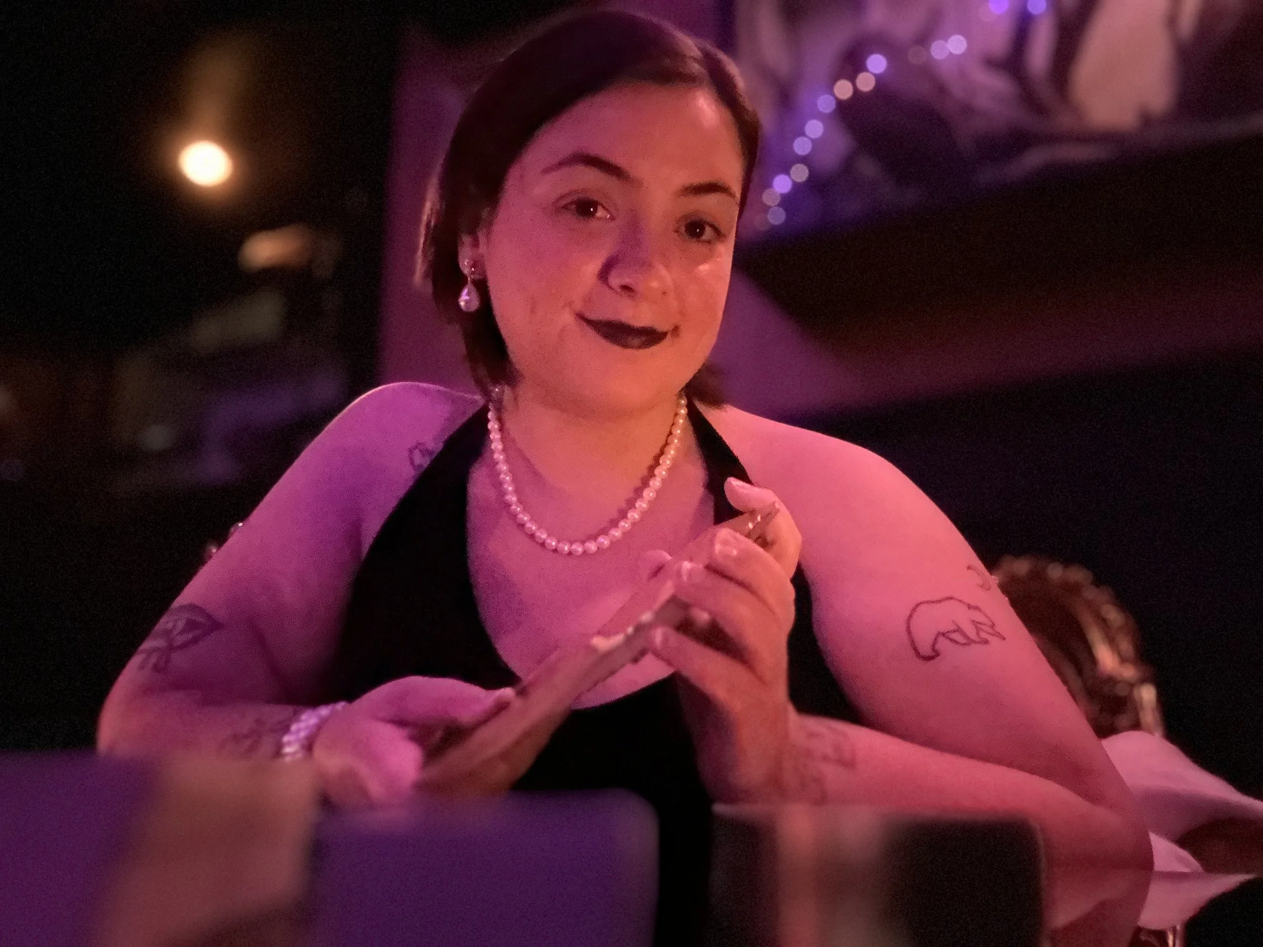 Smiling woman in a pearl necklace and dangling earrings posing with tarot cards under purple and pink evening lights.