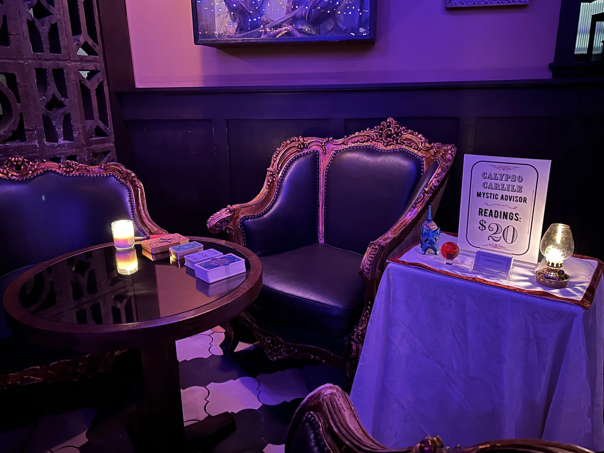 Purple-lit tarot reading room with ornate chairs, tarot cards, candles, crystal ball, and a $20 psychic reading sign for Calypso Carlile.