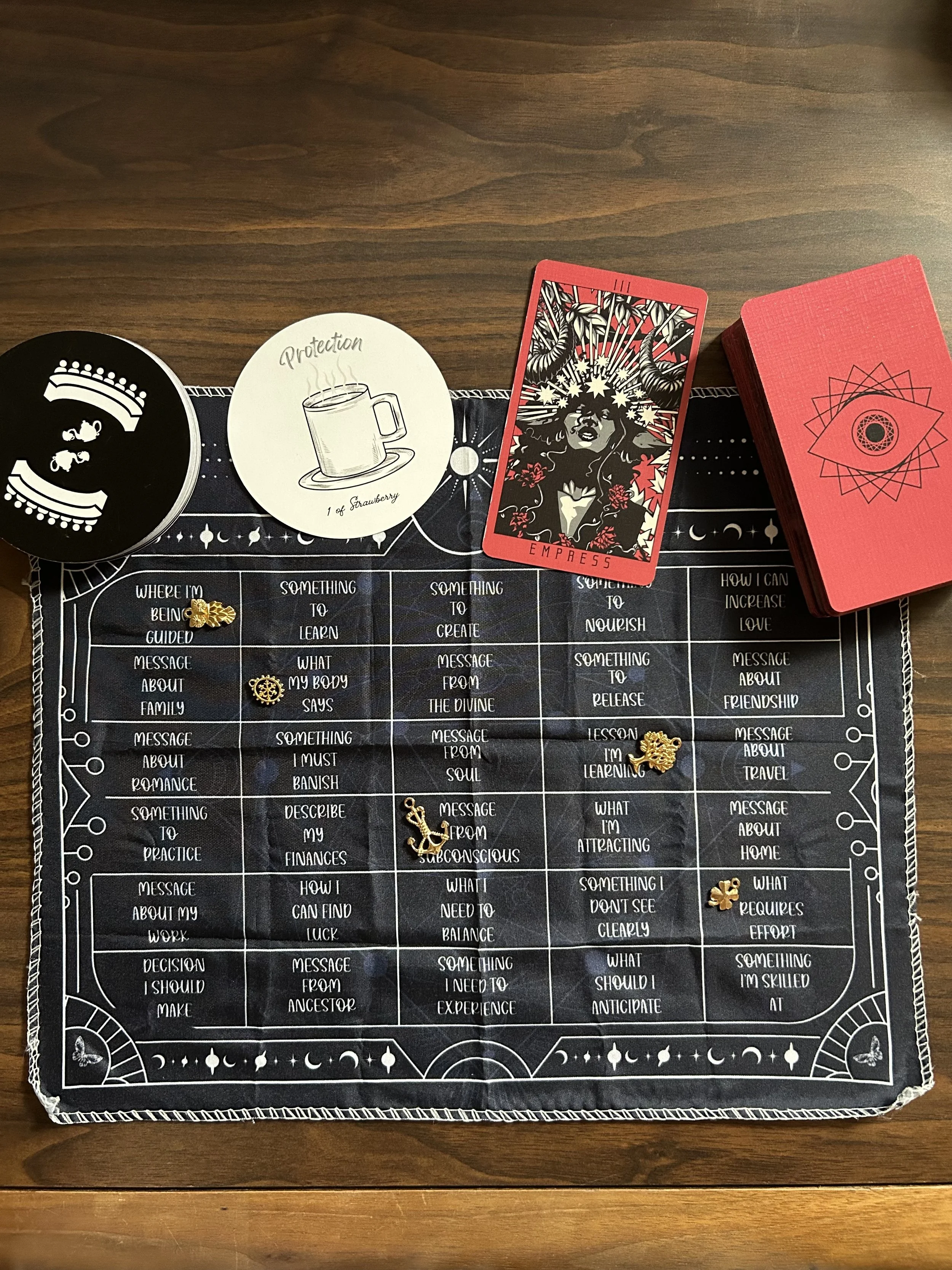 Tarot reading setup on a wooden table with a black divination cloth, gold charms, circular oracle cards, a red Empress tarot card, and a red card deck.