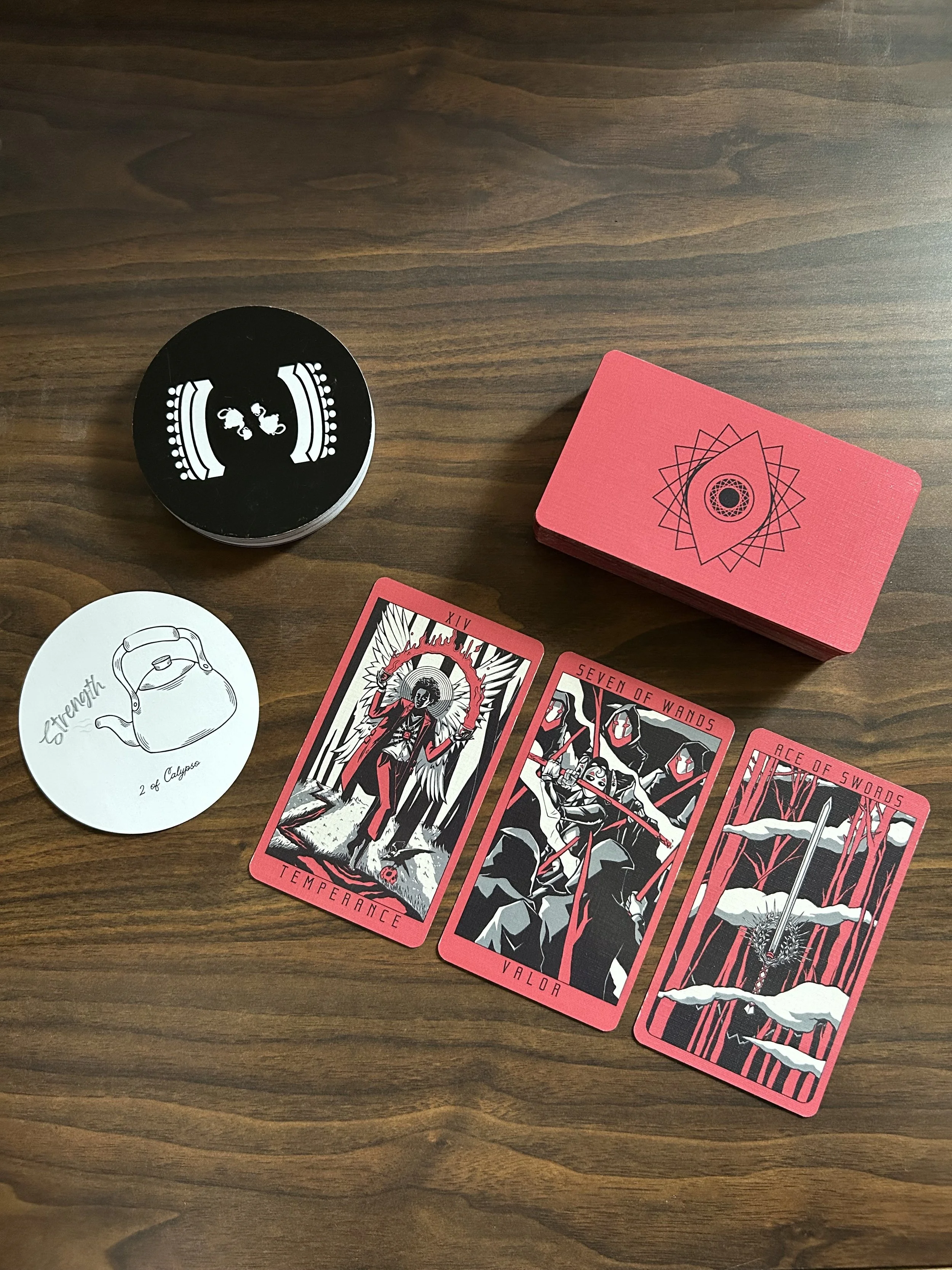 Tarot cards and deck boxes arranged on a dark wooden table, including red-and-black illustrated cards and a black circular card deck.