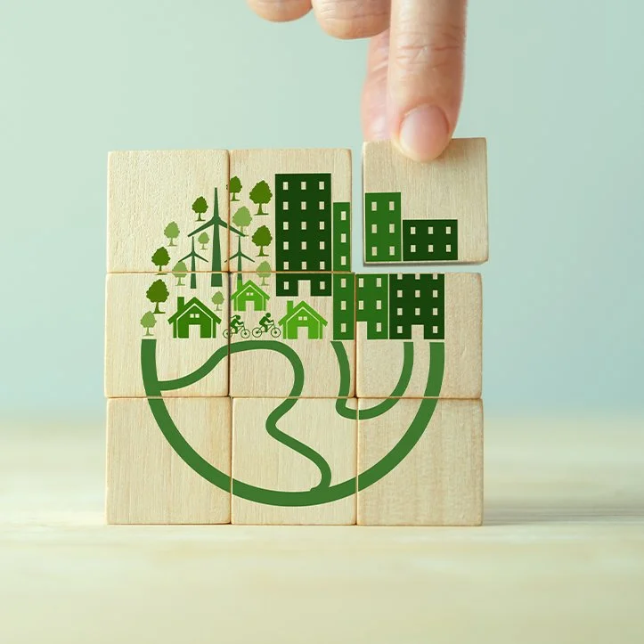 Hand placing a wooden block into a sustainable green city puzzle with trees, buildings, homes, bicycles, wind turbines, and winding paths
