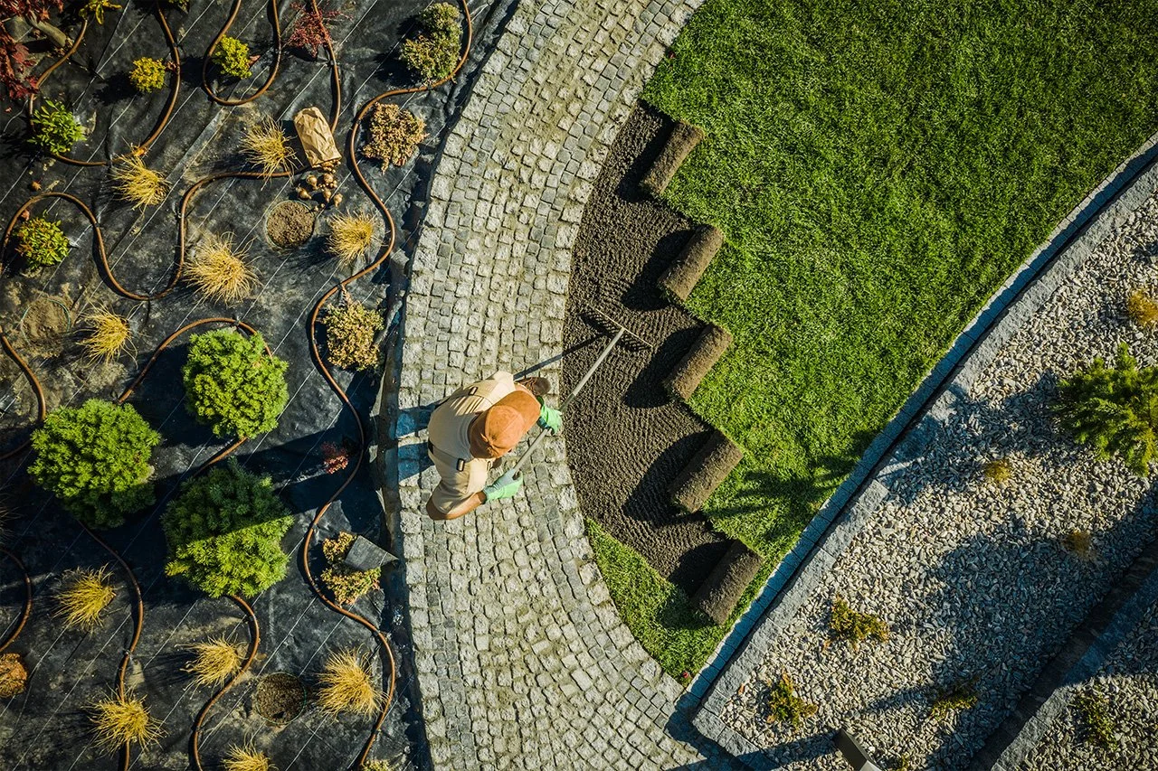 A landscaper installs rolls of lawn beside a cobblestone path, planted garden bed, and decorative gravel landscaping.