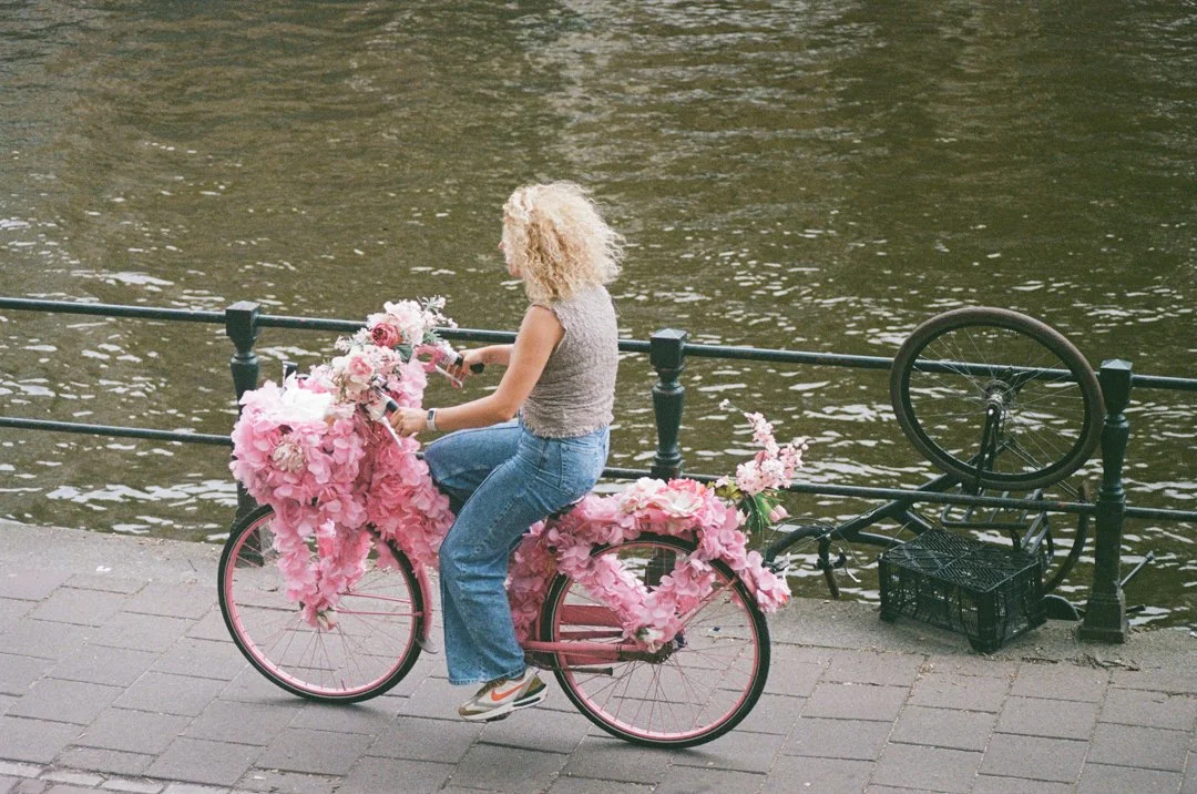 Woman riding a pink bicycle decorated with flowers along a riverside promenade while a black bicycle is about to fall.