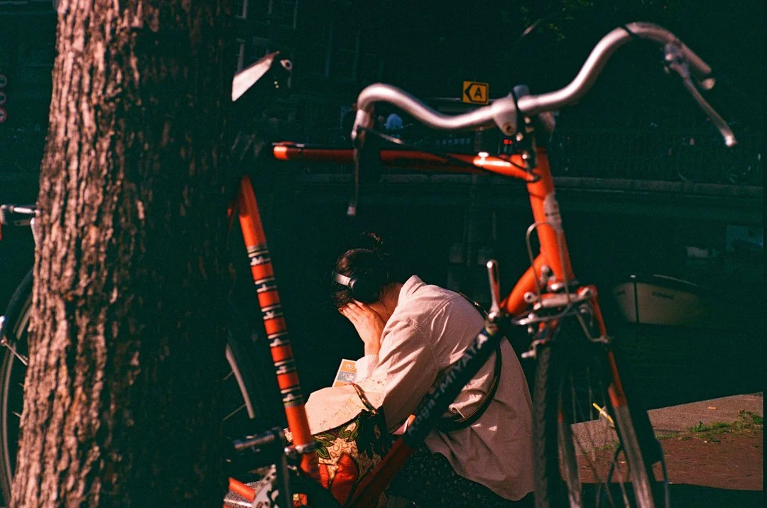 Person wearing headphones crouches beside an orange bicycle locked to a tree on a city street.