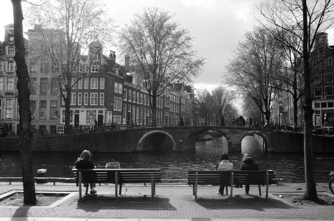 Black-and-white view of people sitting on benches beside an Amsterdam canal, with arched stone bridges, bicycles, trees, and historic canal houses.