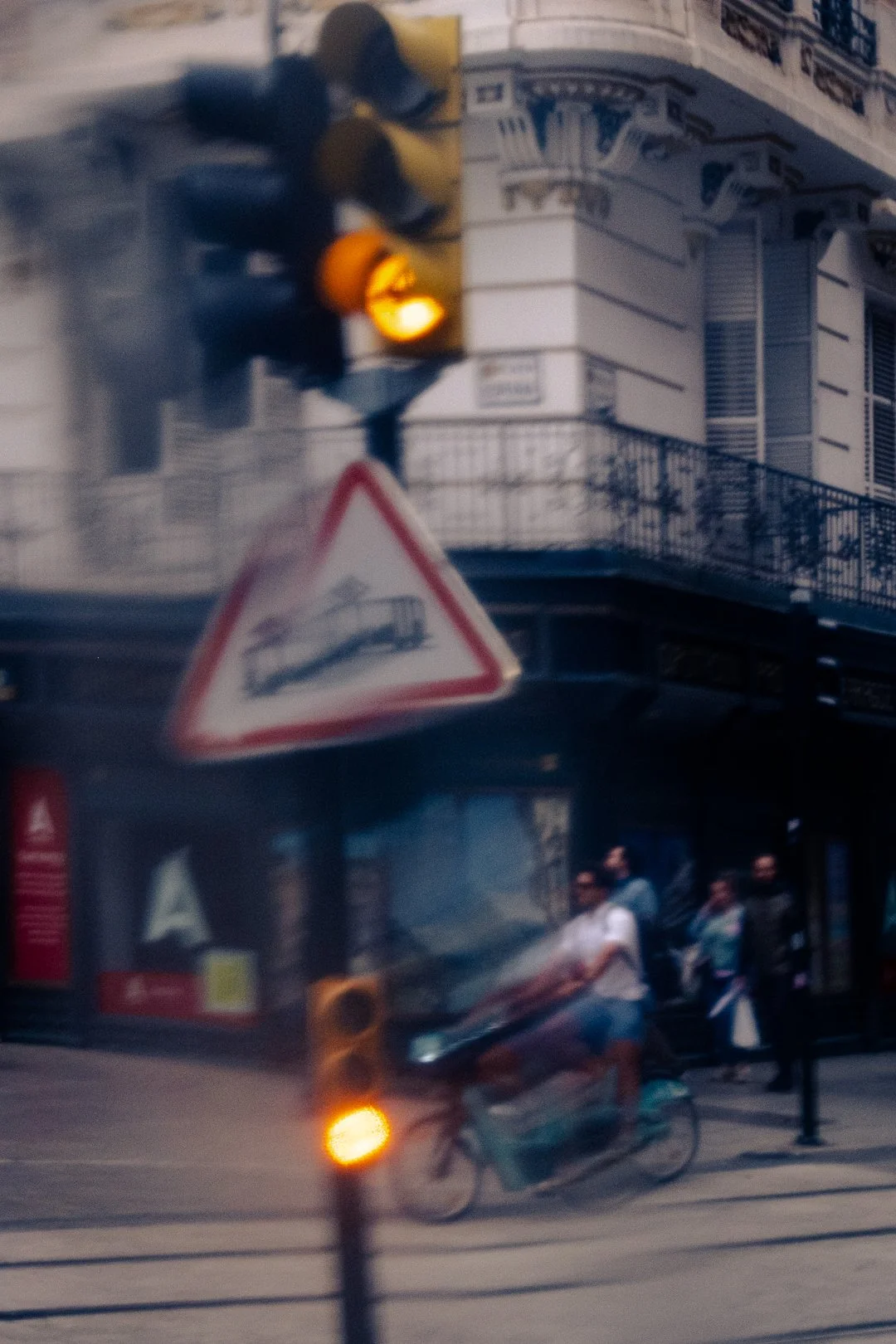 Blurred city street scene with a delivery cyclist, tram tracks, traffic lights, and a triangular roadwork warning sign.