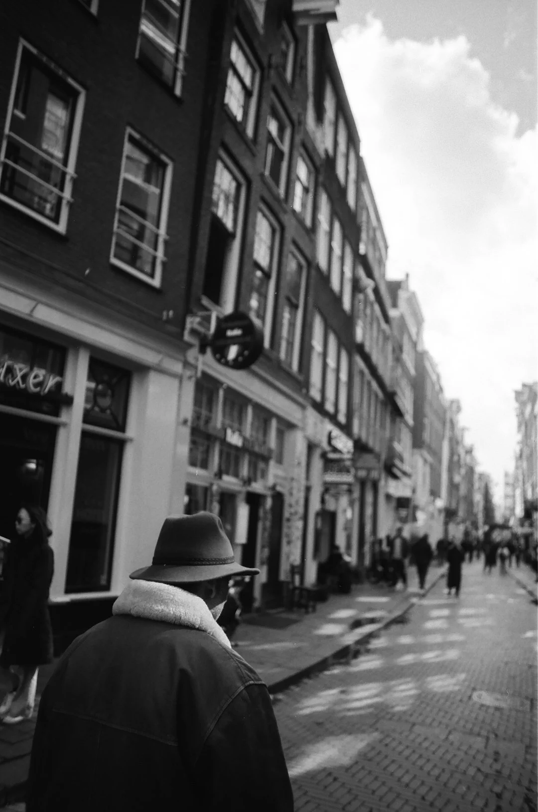 Black-and-white street scene with a person in a wide-brimmed hat walking past historic brick buildings and storefronts.