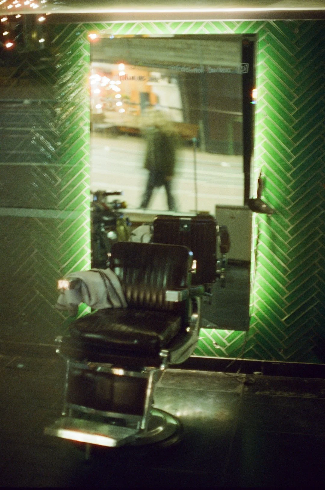 Vintage black barber chair reflected in a large mirror, framed by green chevron-patterned tiles.