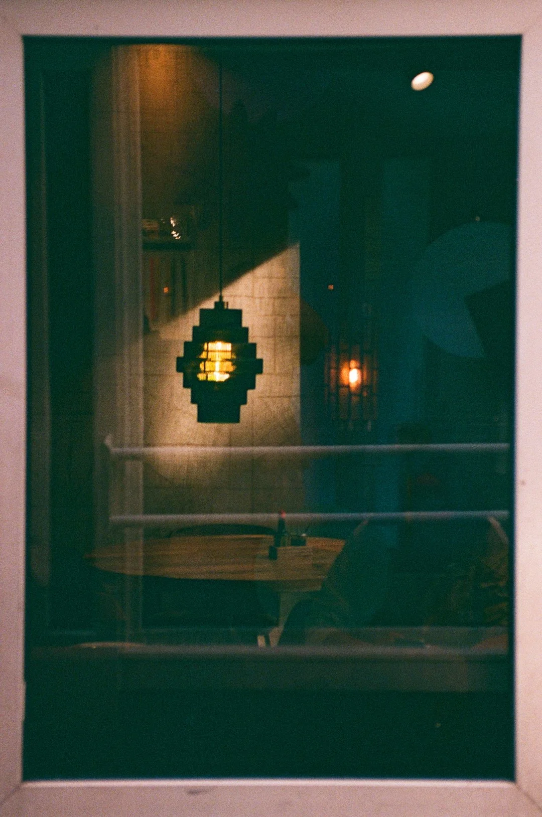 Dimly lit interior reflected in a window, featuring a geometric hanging lantern, wooden table, and warm wall lights.